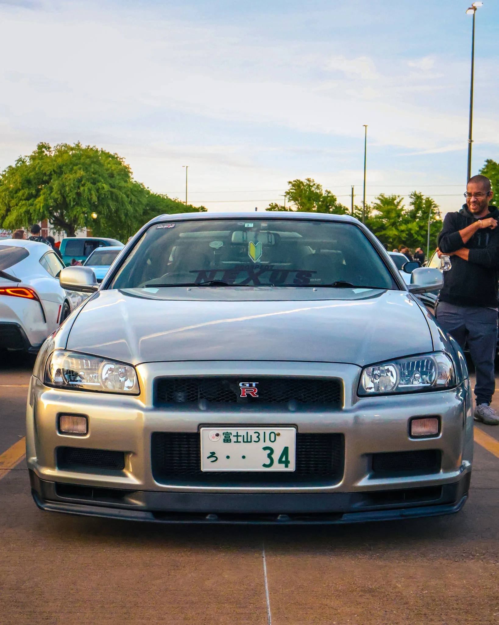 Front view of a silver Nissan GT-R sports car parked in a lot during daytime, with a man standing beside it holding a water bottle and smiling.