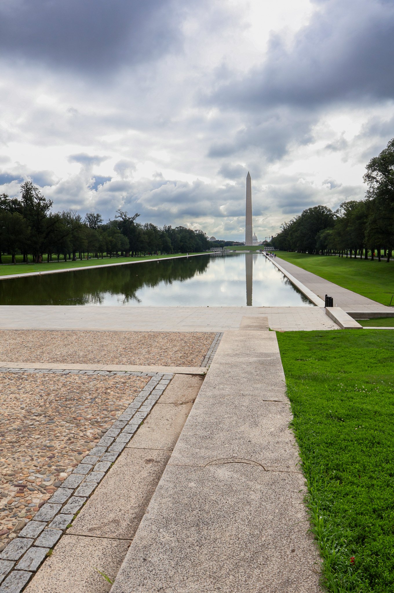Washington Monument reflected in the reflecting pool on a cloudy day, surrounded by green grass and trees.