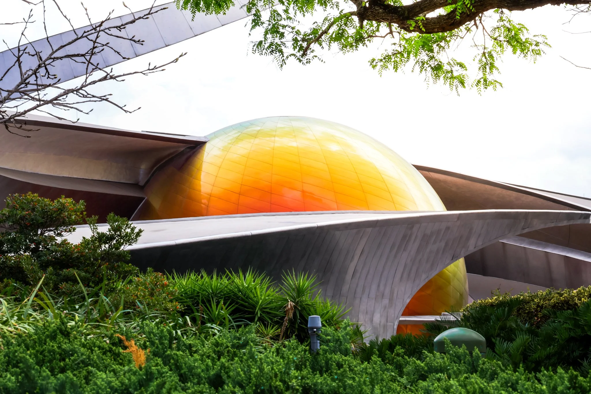 Unique modern building with a large orange, yellow, and reflective dome, surrounded by lush green plants and trees, with a partly cloudy sky in the background.