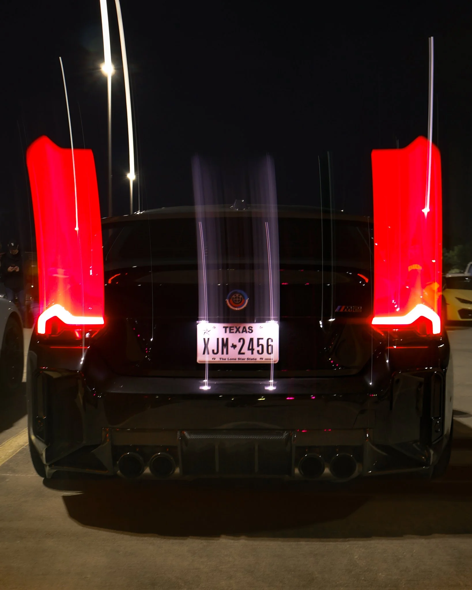Rear view of a black sports car with illuminated vertical taillights, Texas license plate reading 'JM-2456', parked at night in a parking lot.