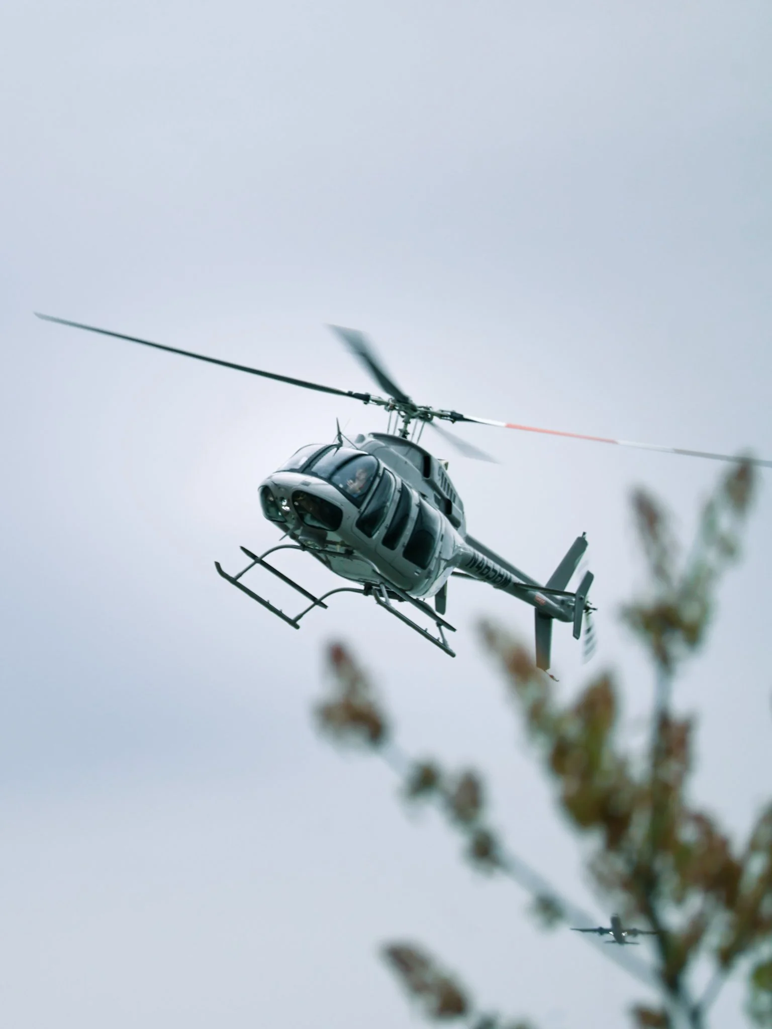 A gray helicopter flying in the sky with another smaller helicopter visible in the background and some blurred tree branches in the foreground.