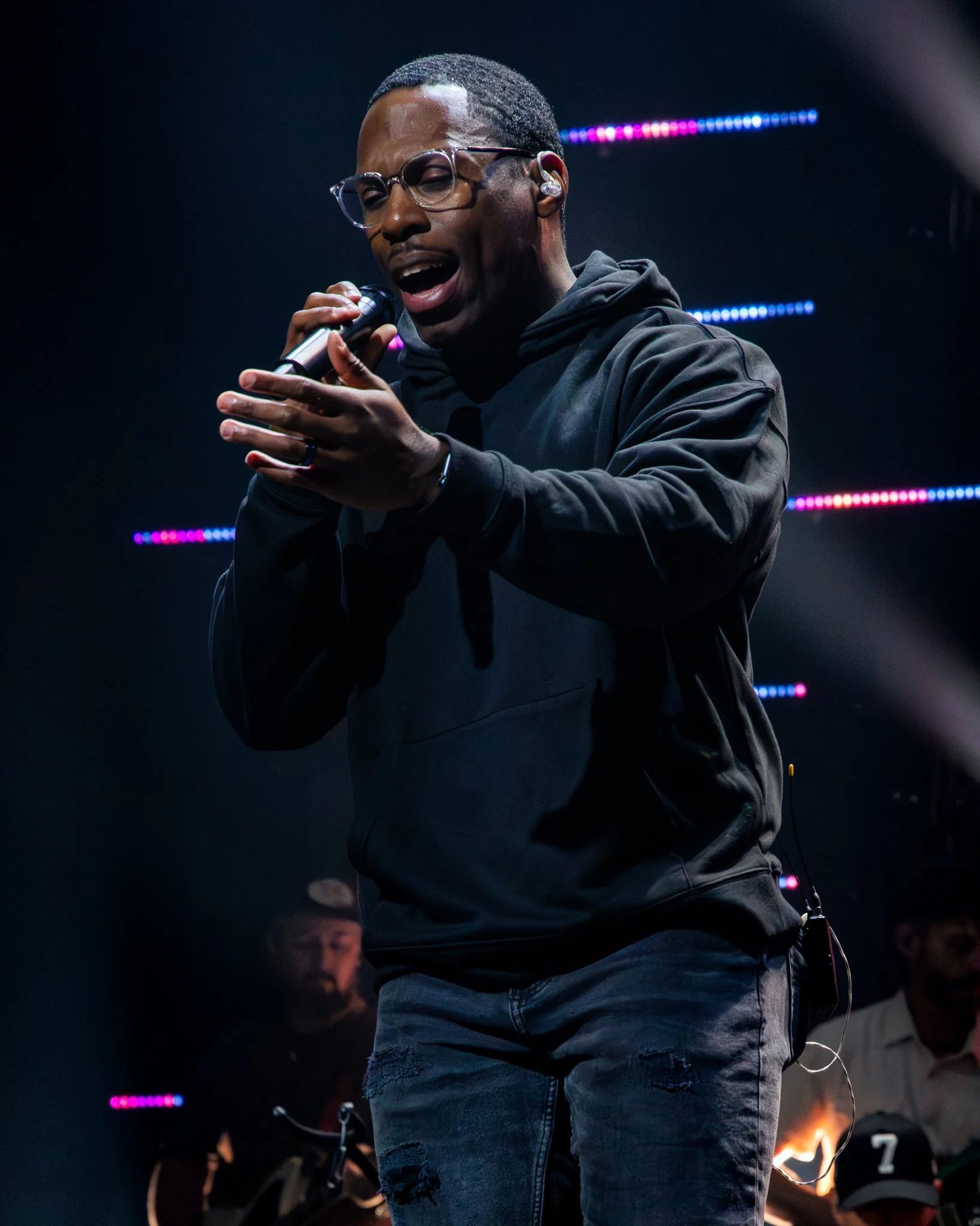 A man singing into a microphone on stage, wearing glasses and a black hoodie, with colorful stage lights in the background.