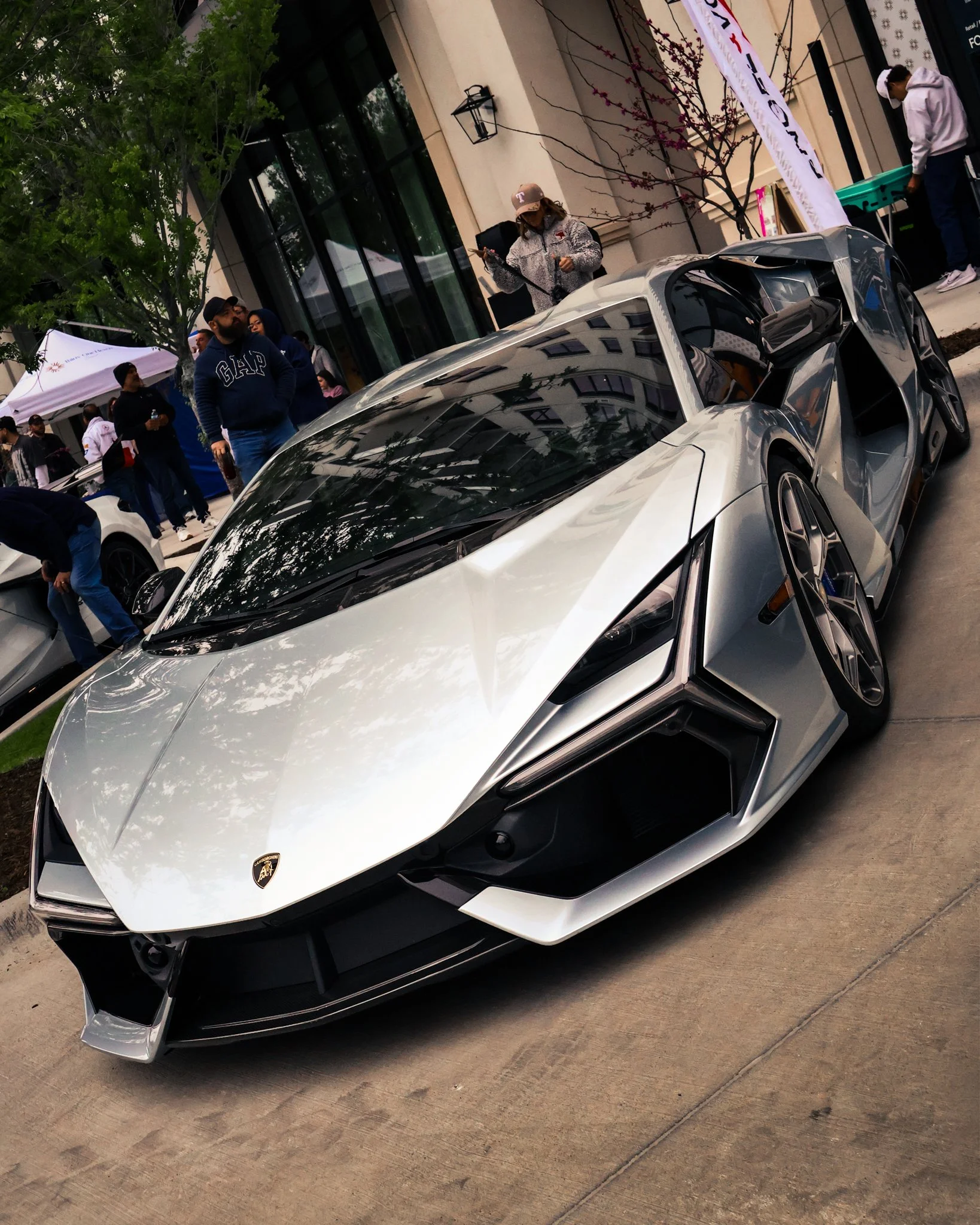 A silver Lamborghini sports car displayed outdoors at an event with people and tents in the background.