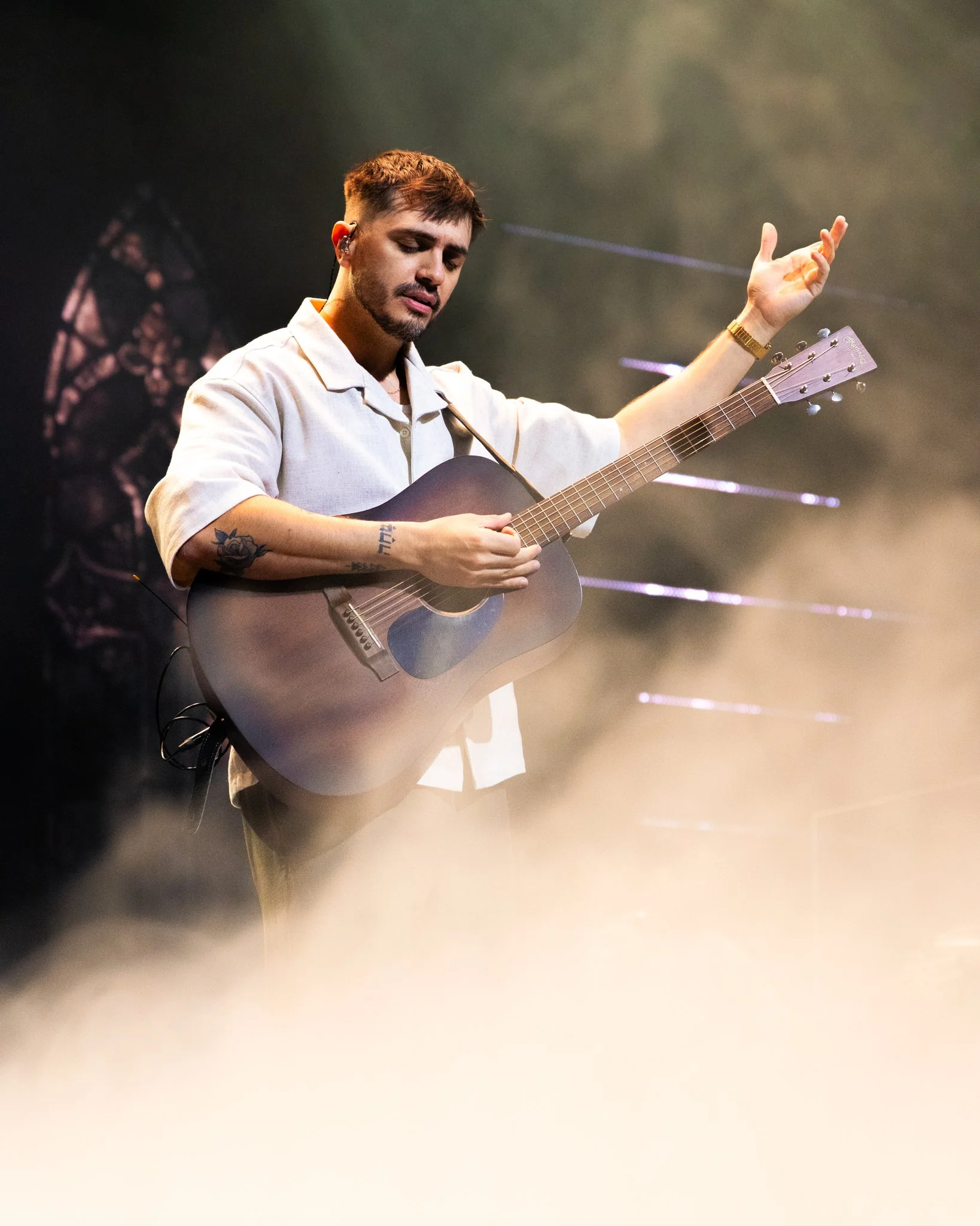 A man playing an acoustic guitar on stage with his eyes closed, wearing a white shirt with rolled sleeves, a watch, and tattoos on his arm, with a foggy stage background.