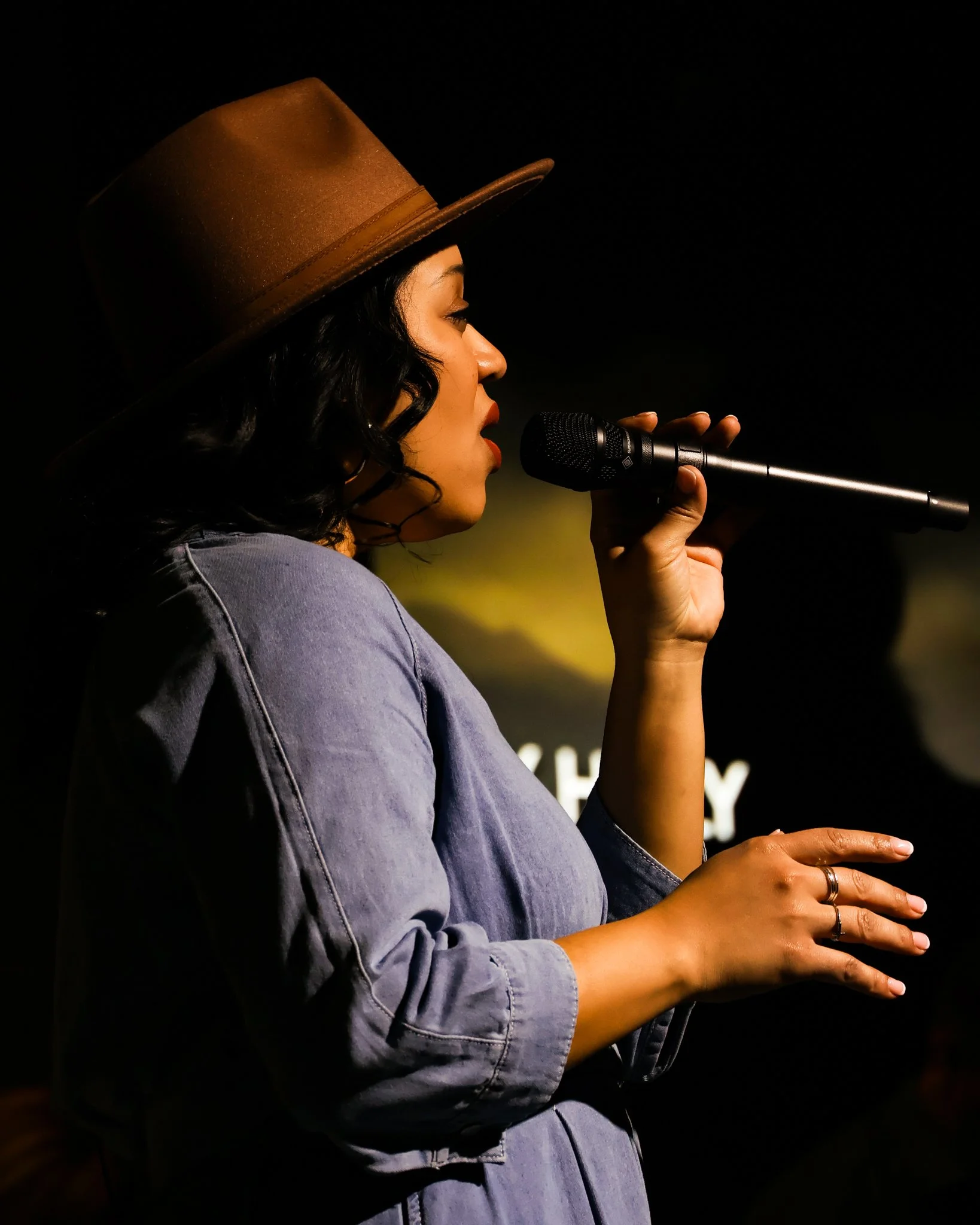A woman with black curly hair, wearing a brown hat and a light denim shirt, holding a microphone and singing or speaking in a dark indoor setting.