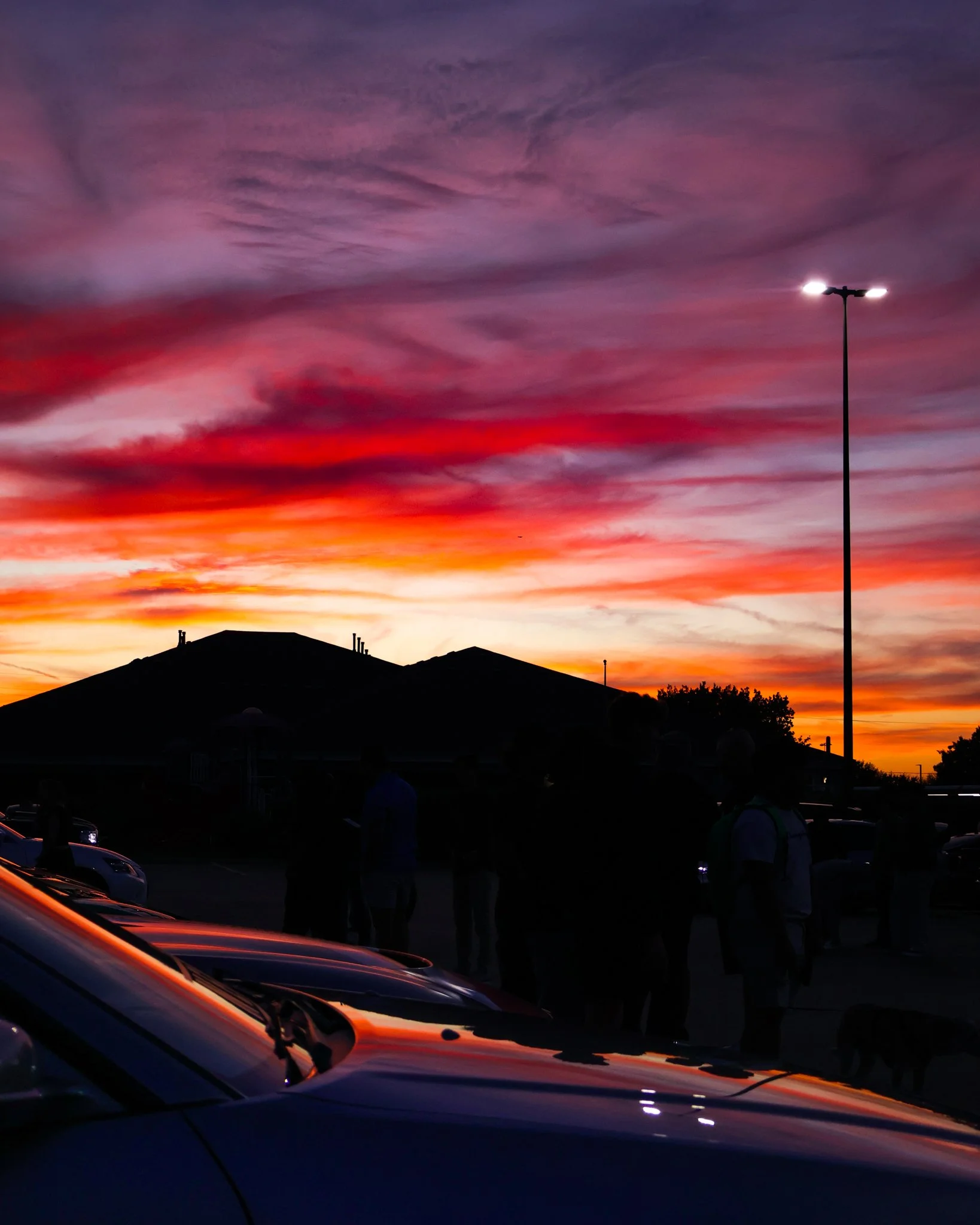 Silhouettes of people and cars against a colorful sunset sky with clouds, a streetlight, and a hill in the background.