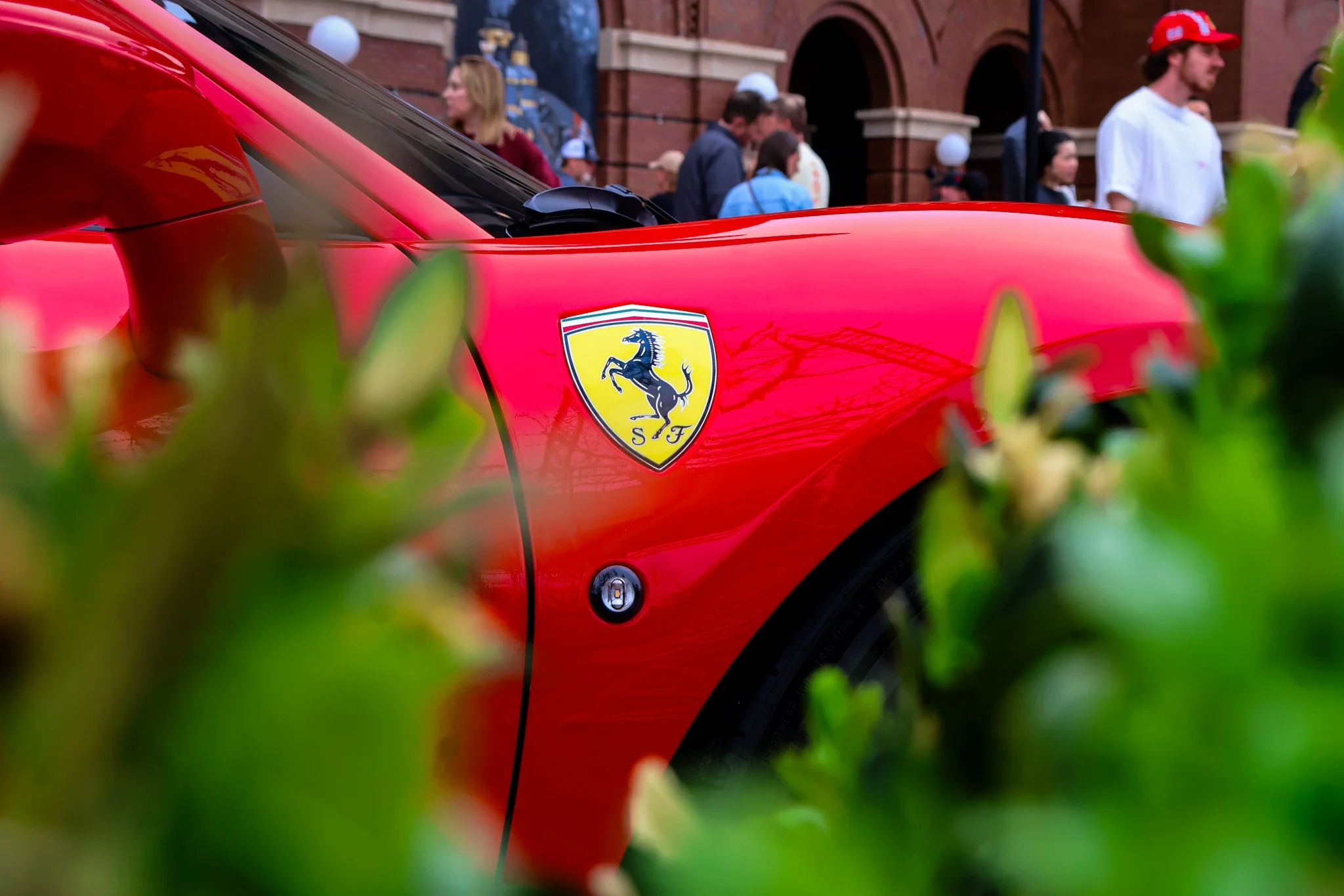 Close-up of a red Ferrari sports car showing the Ferrari emblem, with a crowd of people in the background and some green foliage in the foreground.