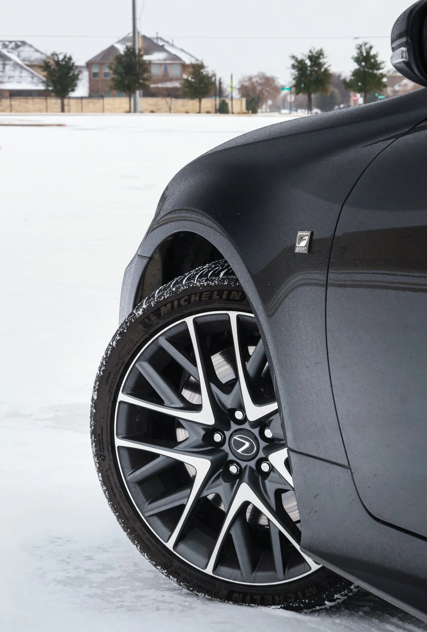 Close-up of a black car tire with snow on the edge of the rim on a snowy landscape, with houses and trees in the background.
