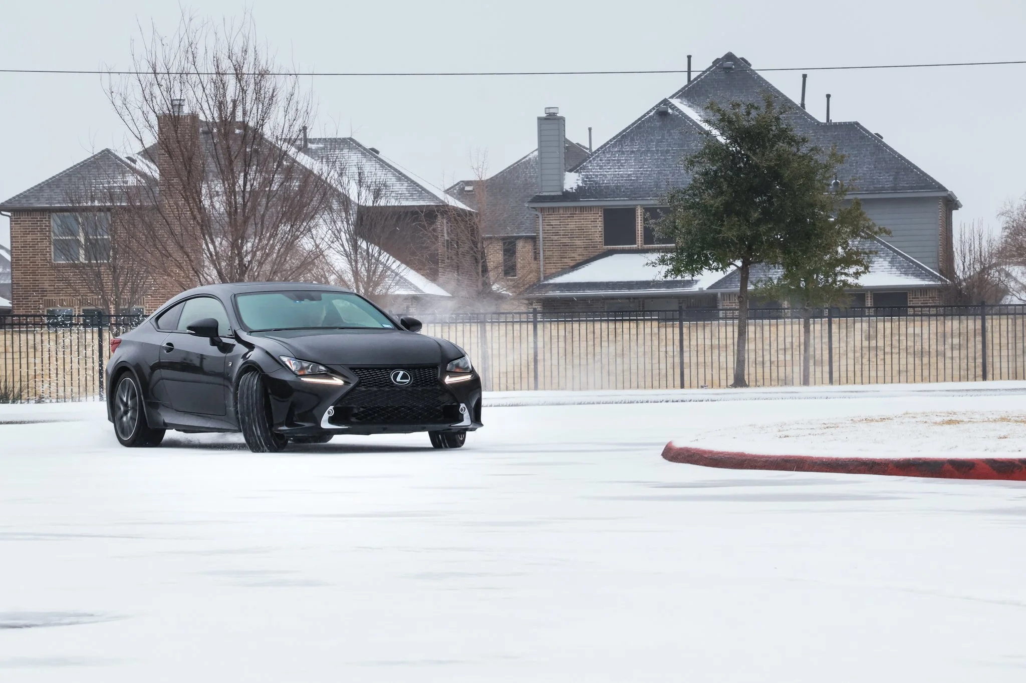 A black Lexus coupe driving on a snow-covered parking lot with a red curb and a tree in the background, houses and leafless trees also visible.