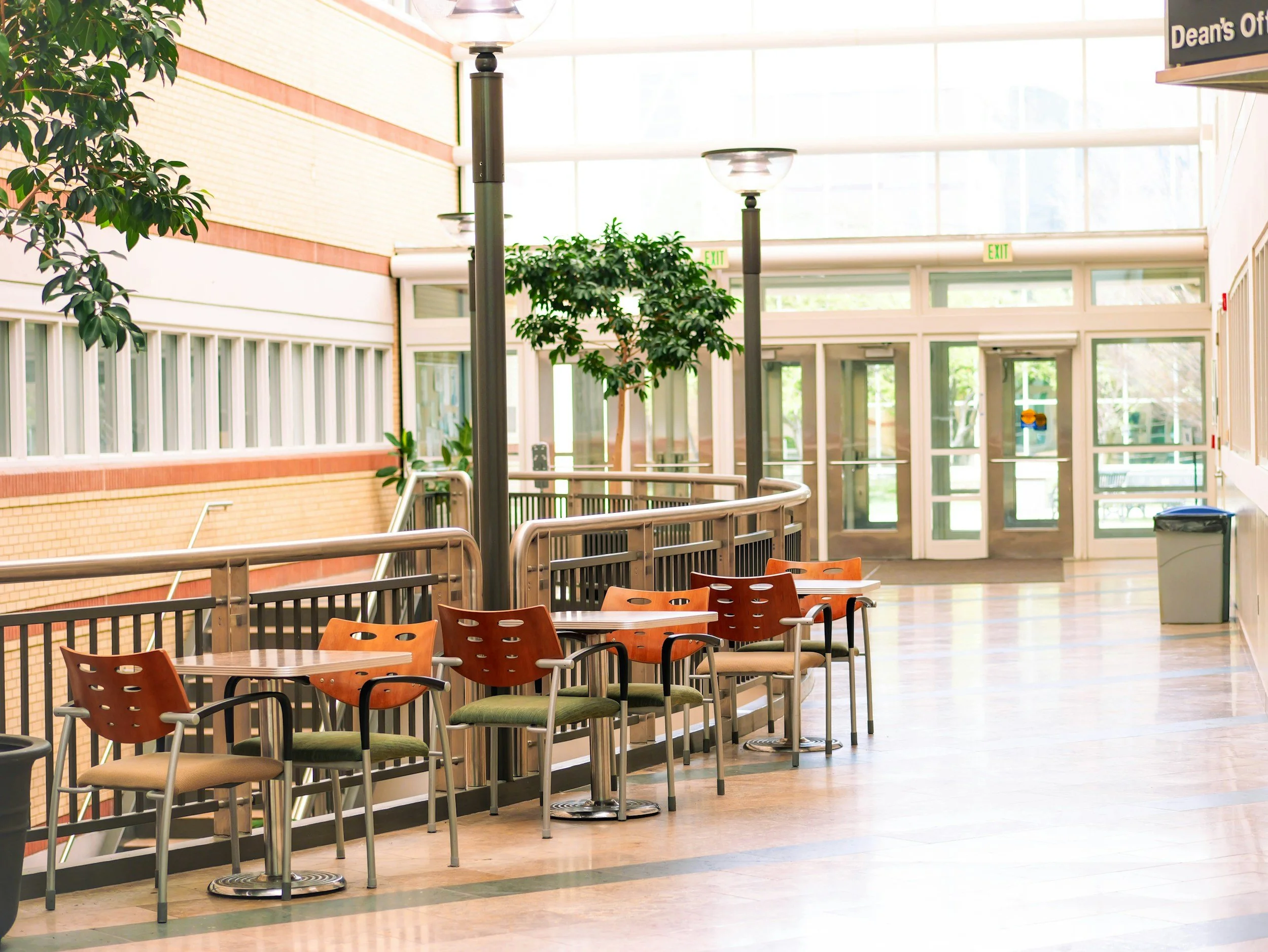 Empty indoor waiting area with chairs, small tables, potted plants, and large windows in a modern public building.