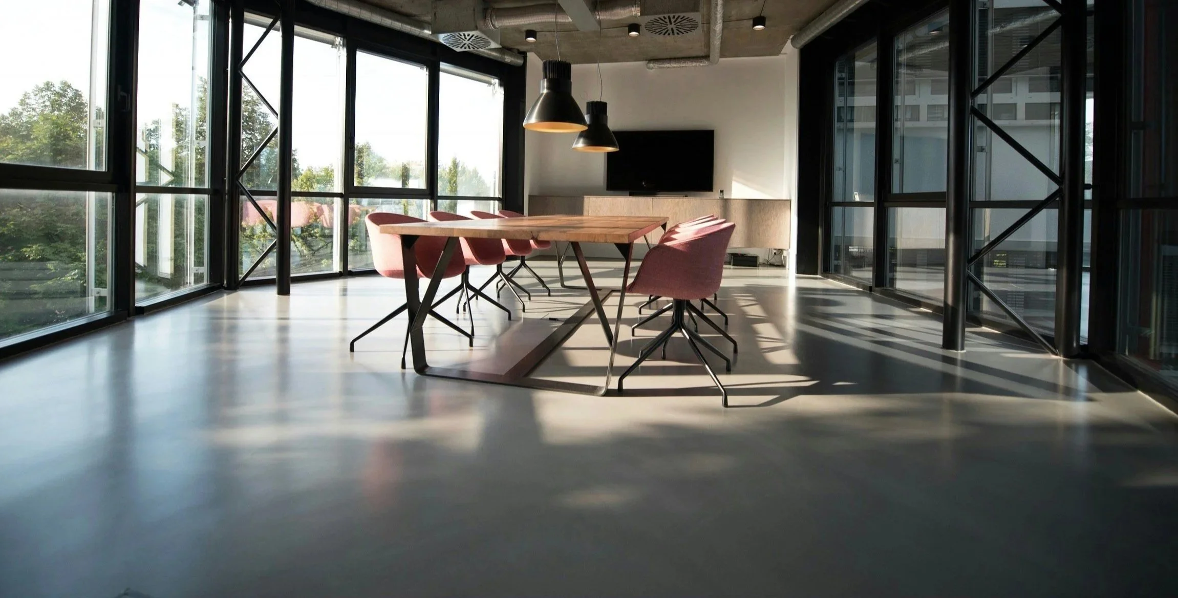Empty modern conference room with a wooden table, six pink chairs, large windows with black frames, a flat-screen TV on the wall, and black hanging light fixtures.