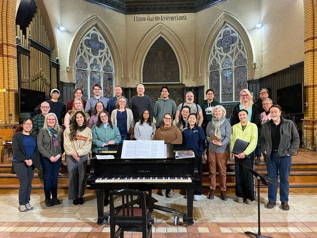 Group of our choristers posing behind a grand piano inside a church, with stained glass windows and brick walls.