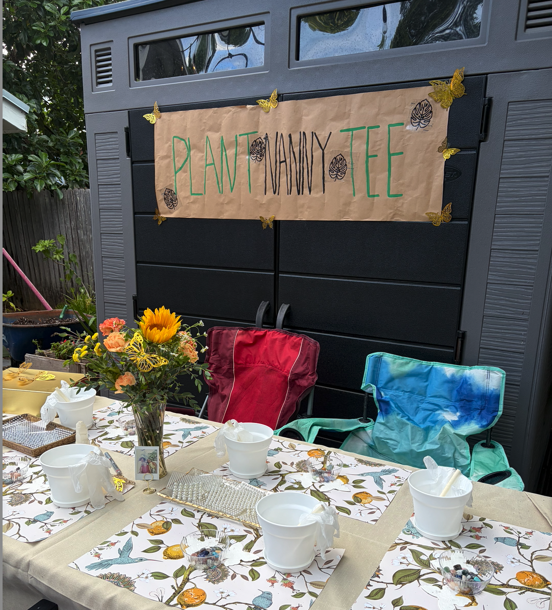Outdoor table decorated with a floral tablecloth, cupcakes in white buckets, and a vase of sunflowers and roses, set for a gathering. A banner reading 'PLANT TEE' with leaf and butterfly decorations hangs on a gray shed in the background, which also 