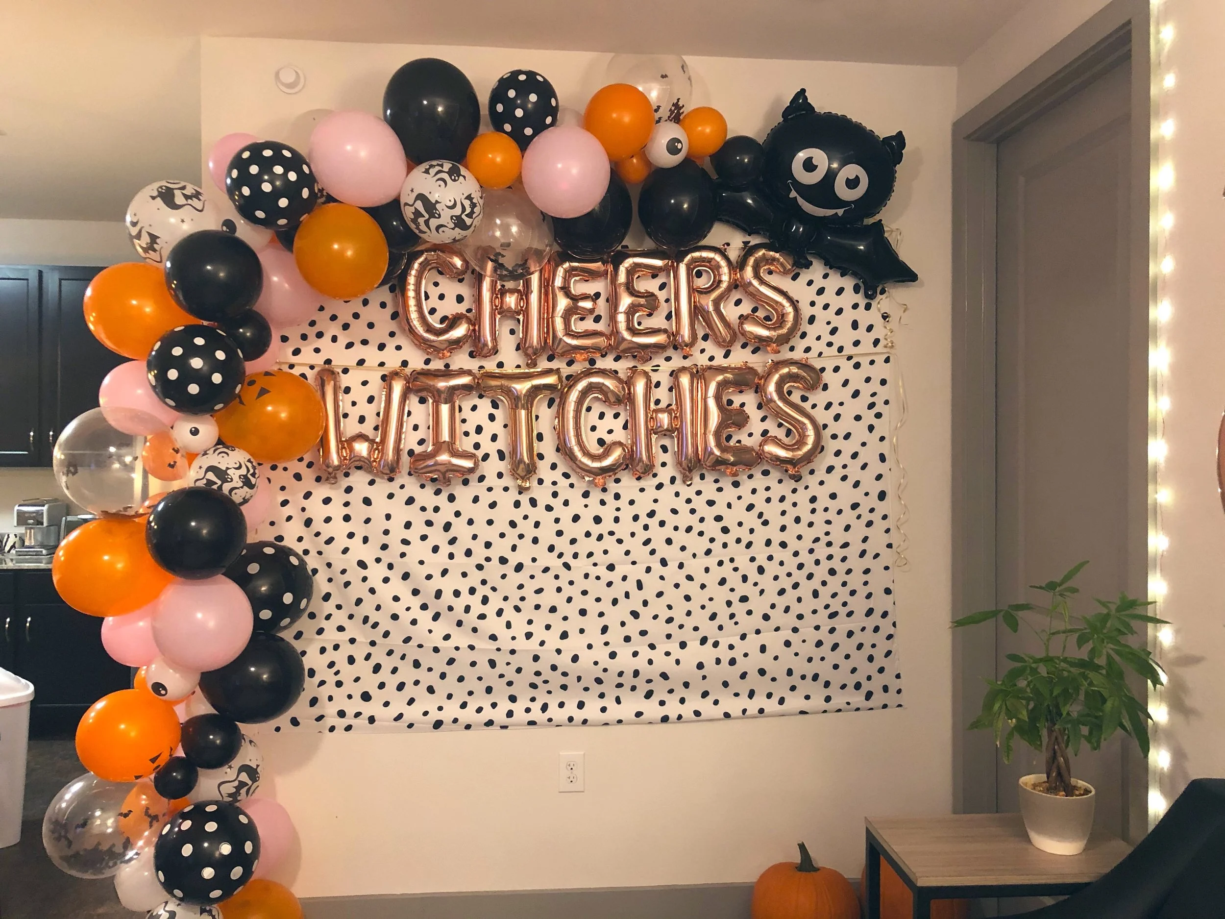 Halloween-themed balloon decoration with black, orange, pink, white, and clear balloons, some with polka dot and spooky face patterns, and a Frankenstein monster-shaped balloon, with metallic balloon letters spelling "CHEERS" on a dot-patterned backd