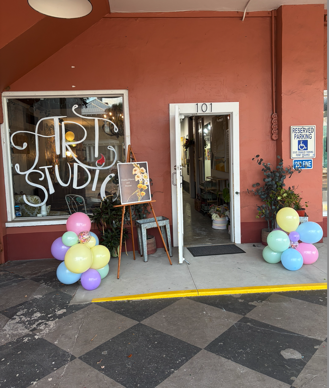 Exterior of a studio with a pink building, balloons in pastel colors on each side of the entrance, and a sign in the window that says 'Vitality Studio'. The door is open, revealing the inside of the studio, with potted plants and artwork visible.