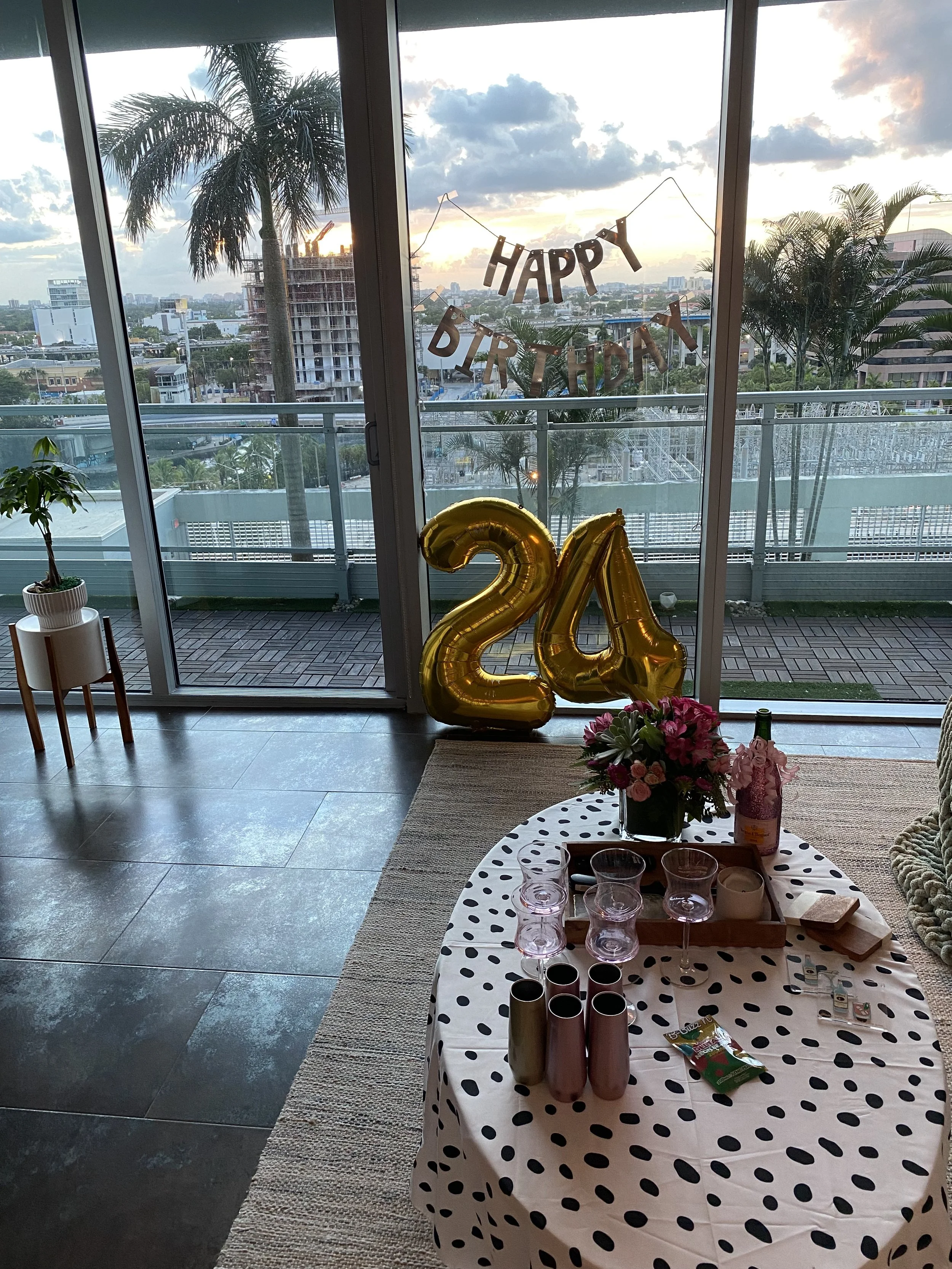 Image of a decorated room with a large window view of a sunset sky and cityscape, a gold '24' balloon, a table with a black polka dot tablecloth, pink flowers, and various party supplies.