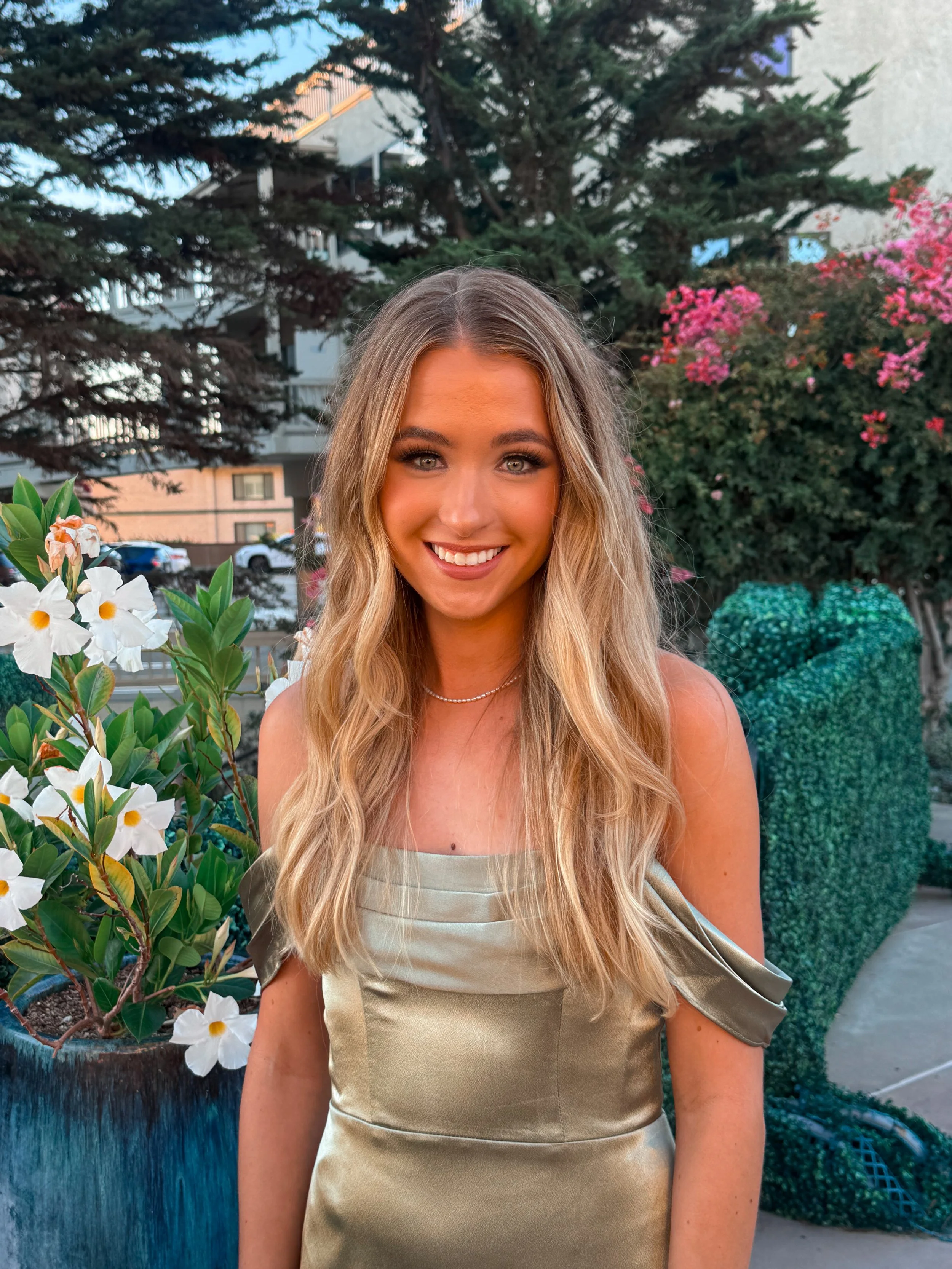 A young woman with long wavy blonde hair and light-colored eyes, wearing a satin champagne-colored dress with off-the-shoulder sleeves, standing outdoors in front of a background with trees, pink flowering bushes, and a building, smiling at the camera.