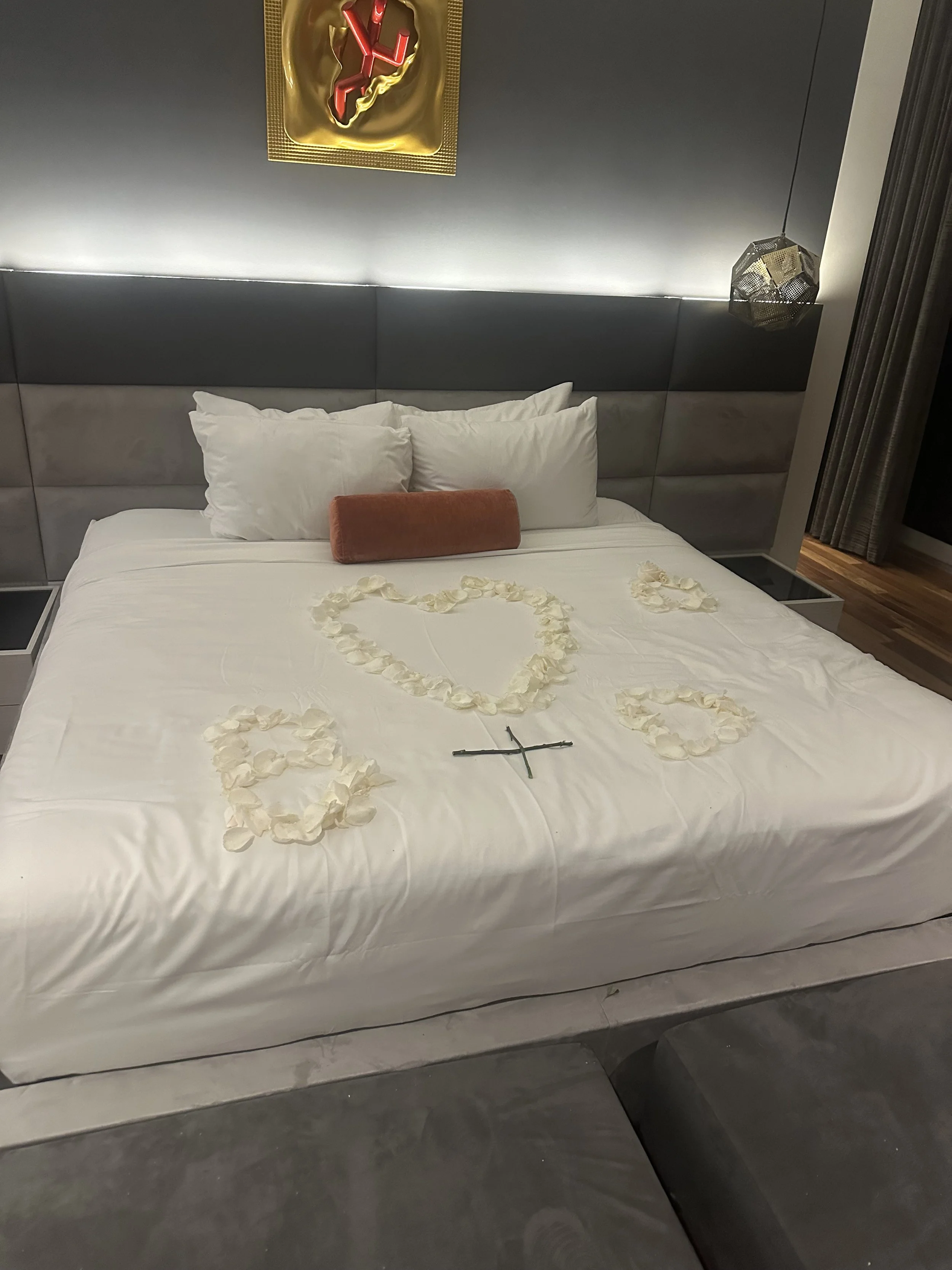 A bed with white sheets and pillows, a heart shape made of rose petals, and a black cross on the bed. There is a gold and red decoration hanging on the wall above the bed and a geometric hanging lamp to the right.