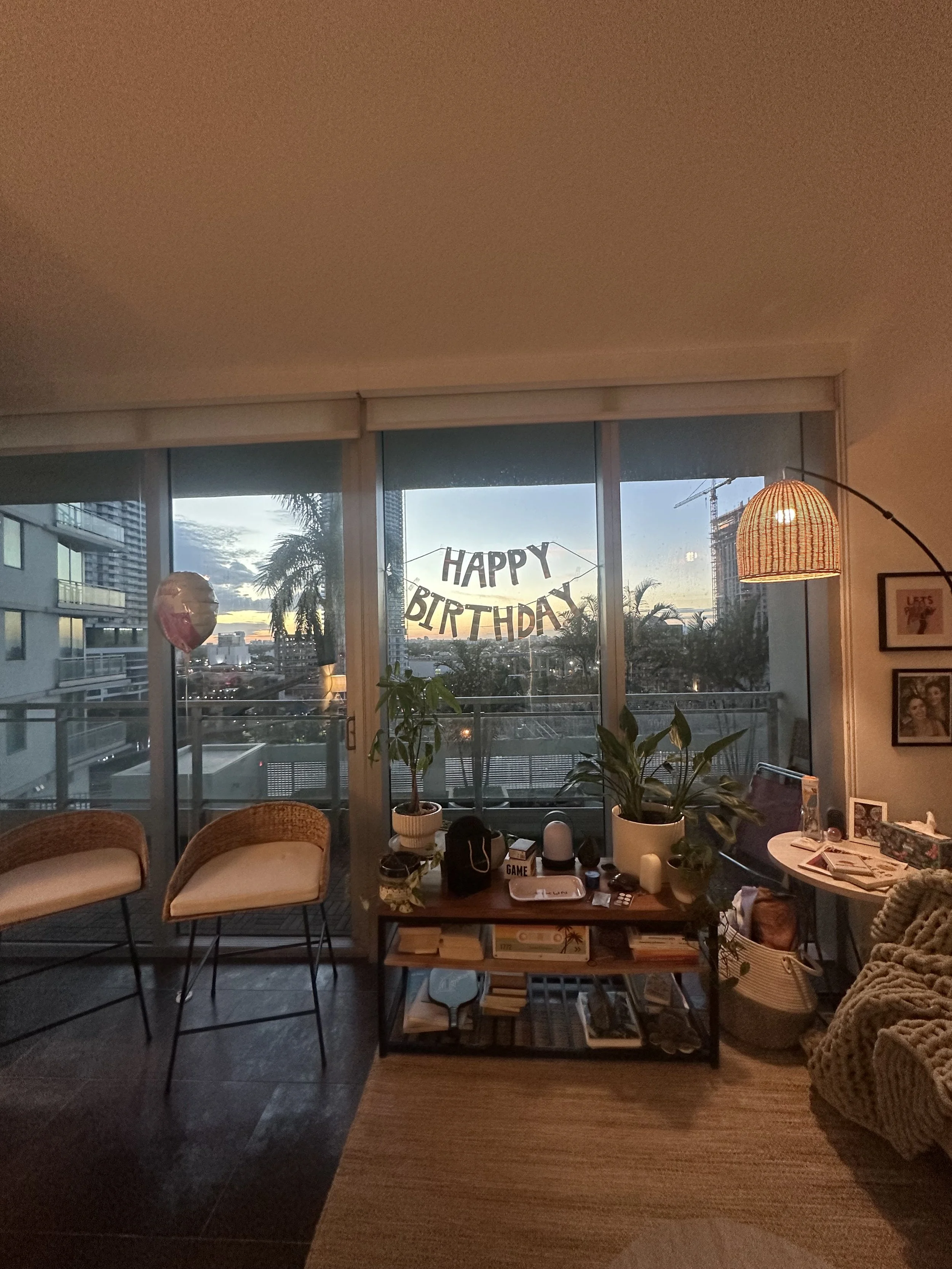 Indoor living space with large sliding glass doors opening to a balcony, decorated with a 'Happy Birthday' banner, interior with chairs, plants, a lamp, framed pictures, and a side table with various items; cityscape and palm trees visible outside.