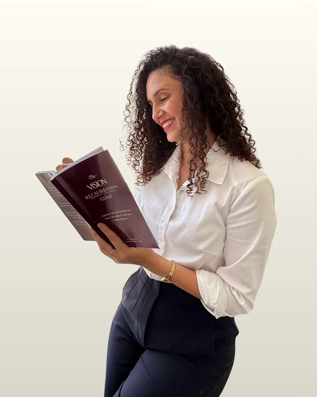 A woman with curly hair, dressed in a white blouse and dark pants, smiling while reading a book titled 'The Vision Recalibration Guide'.