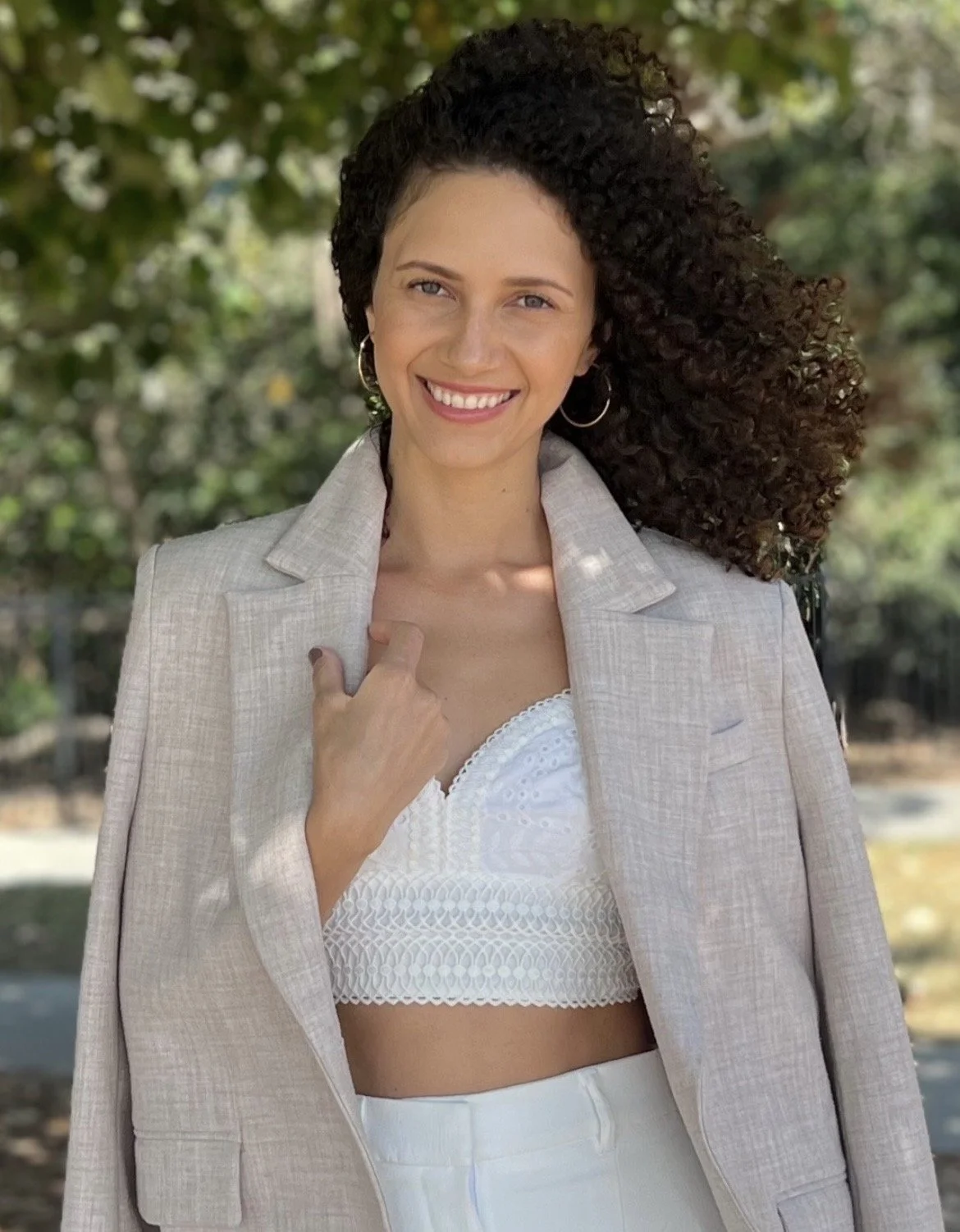 A woman with curly dark hair smiling outdoors, wearing a light-colored blazer over a white lace crop top and white pants.
