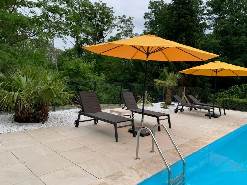 Chaises longues noires avec parasols jaunes autour d'une piscine, avec des plantes et des arbres en arrière-plan.