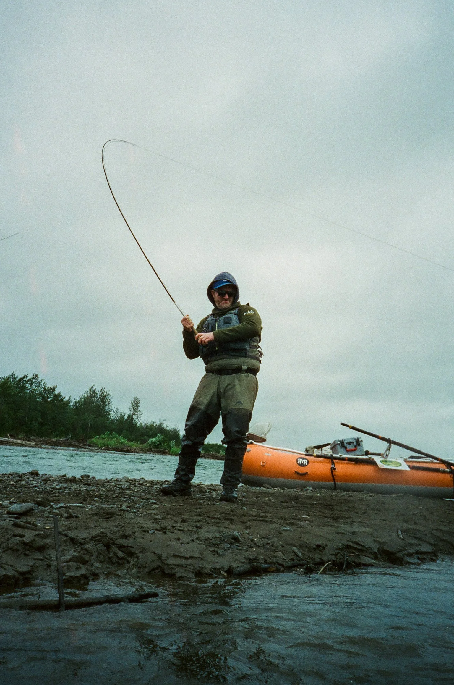 A man wearing a life vest and hat stands on a rocky riverbank holding a fishing rod, with a kayak and fishing gear nearby against a cloudy outdoor backdrop.