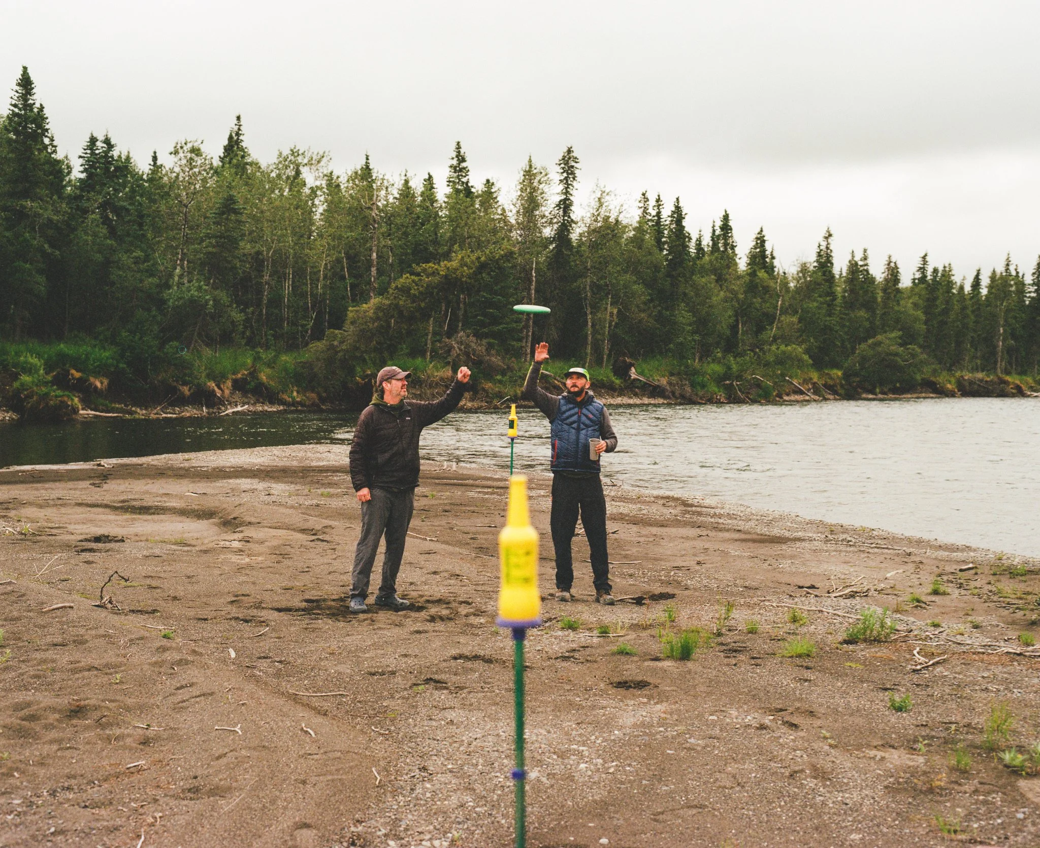 Two men standing on a riverbank, playing with a frisbee, with a forested area in the background.