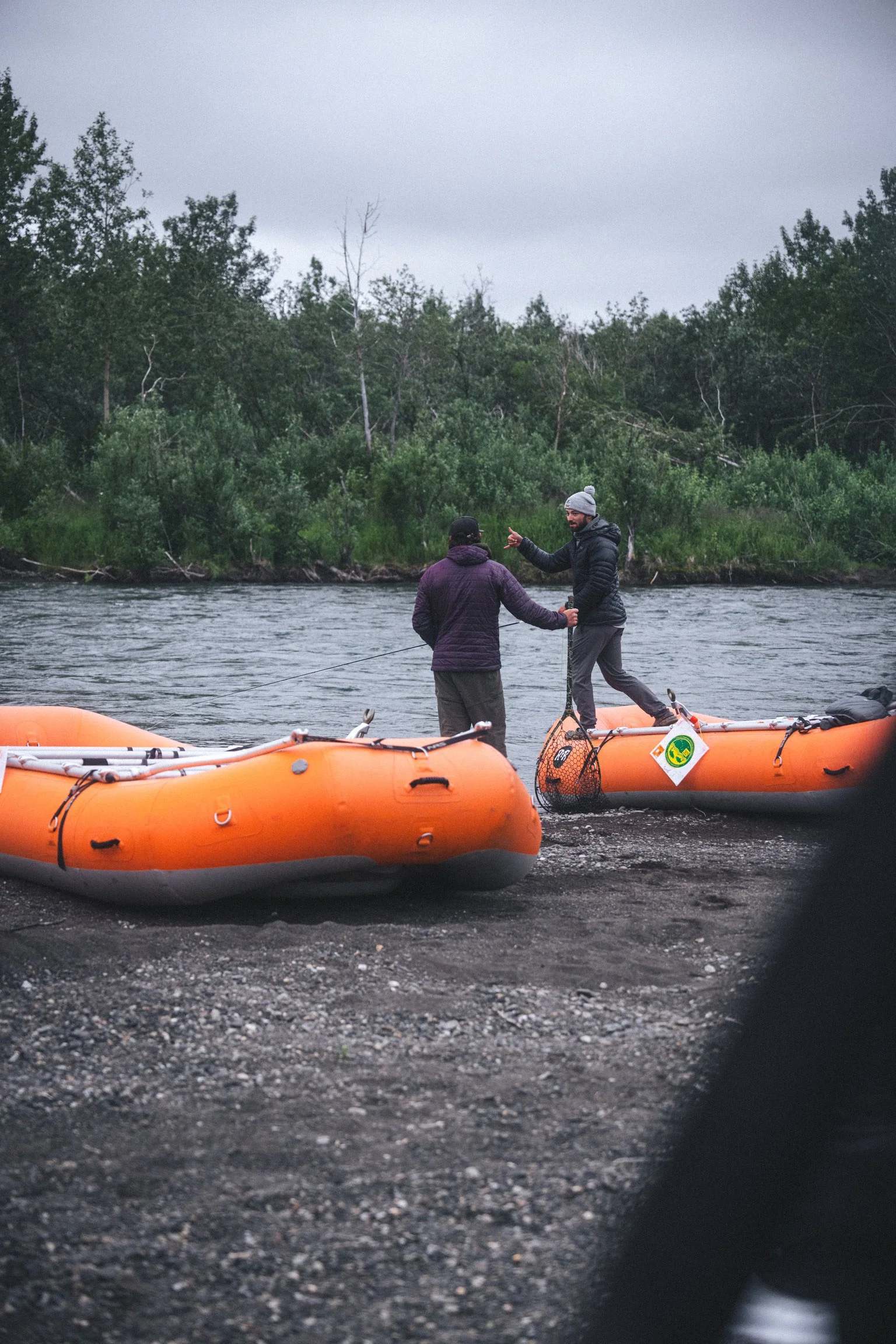 Two men are standing in orange inflatable rafts on a river, with a backdrop of trees and a cloudy sky. One man is gesturing or pointing, and the other is listening. The scene appears to be outdoors in a natural setting, possibly during an overcast da