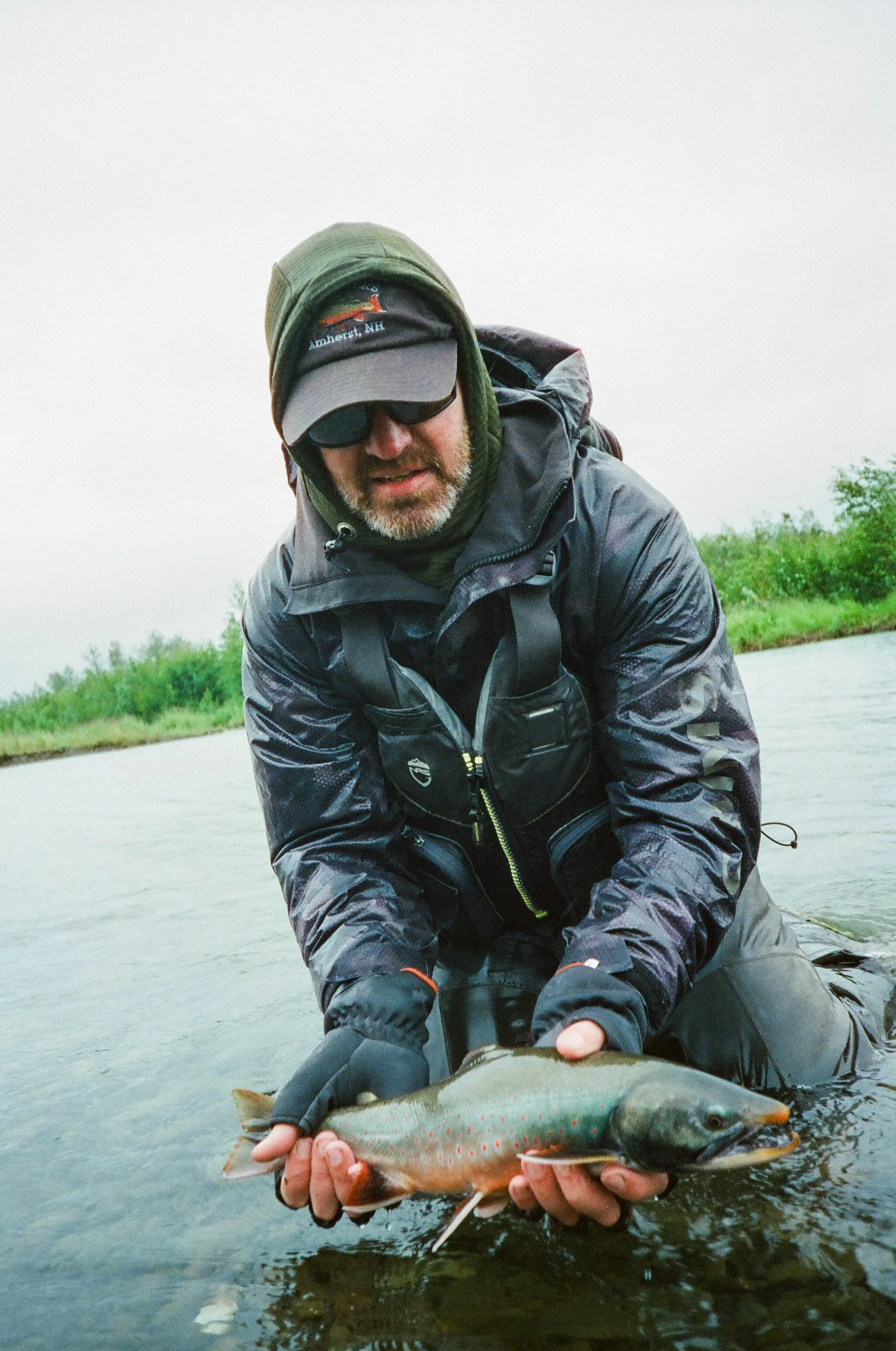 Man wearing outdoor gear and sunglasses holding a large fish over a river or lake with green vegetation in the background.