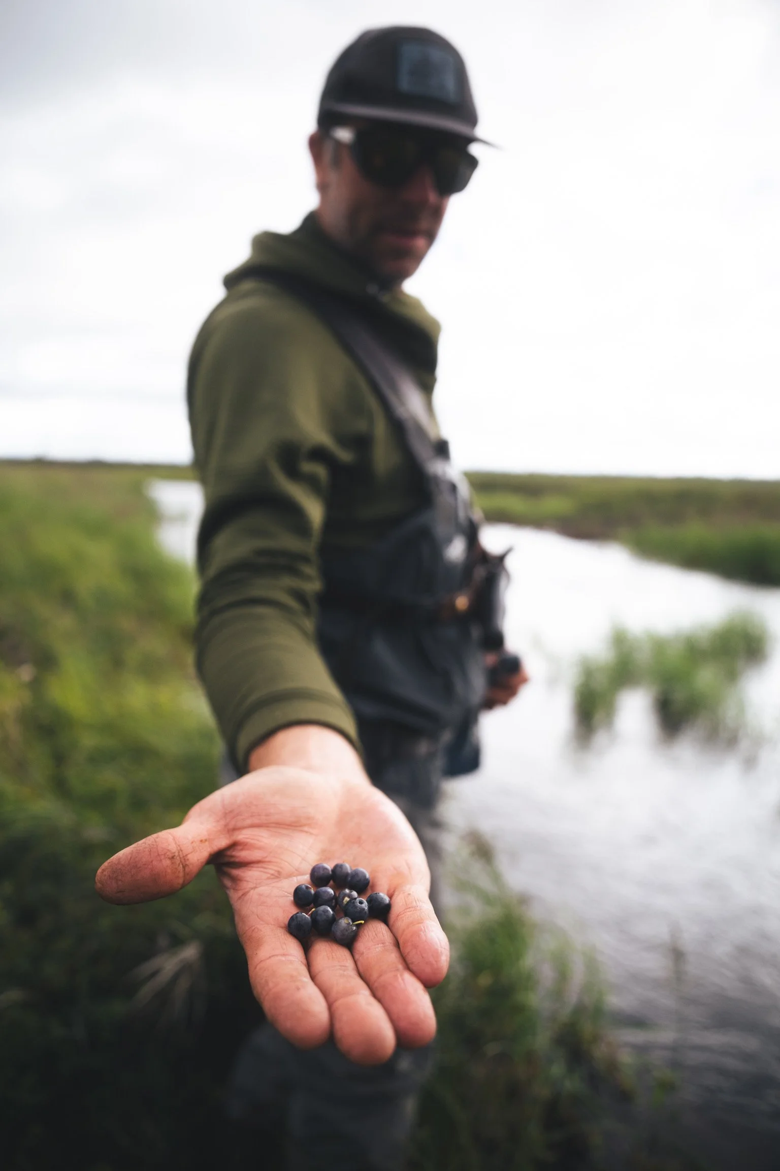 A person holding out their hand with blueberries, standing outdoors by a river with grassy banks under an overcast sky.