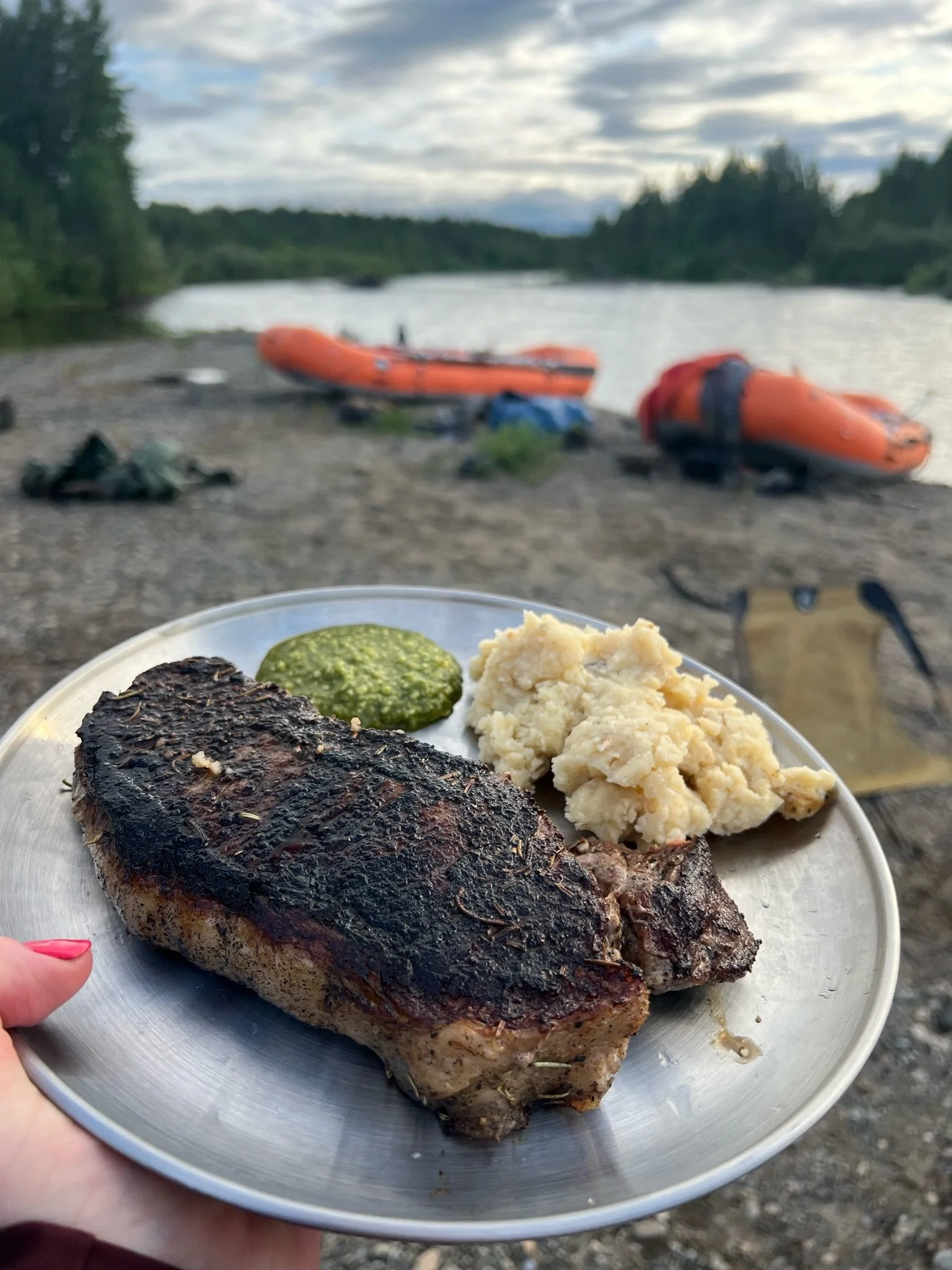 A plate of grilled fish with mashed potatoes and pesto, held outdoors near a river with two orange inflatable boats and camping gear on the shore, under a cloudy sky.
