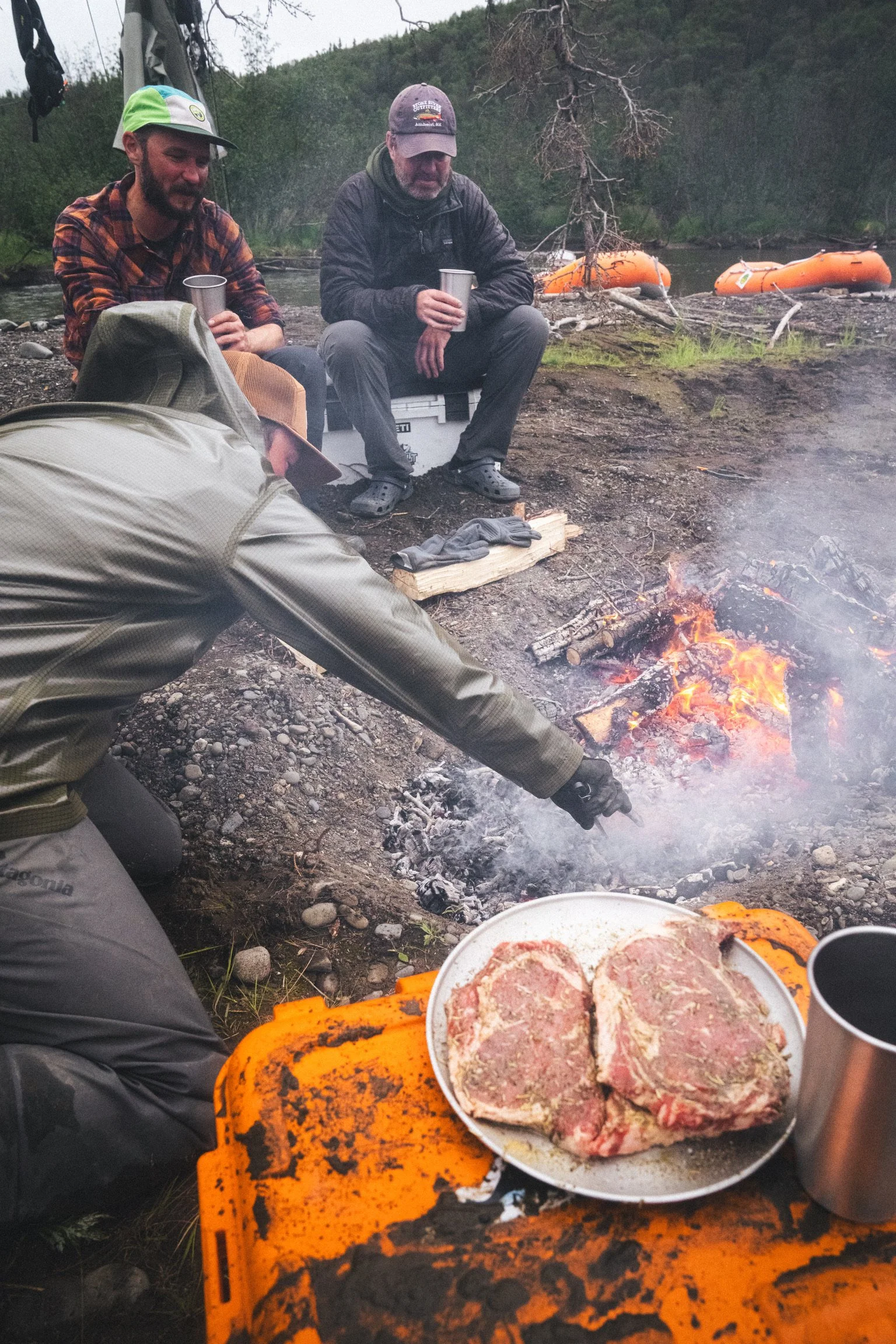 Three men camping outdoors near a river, sitting around a fire with a plate of raw meat and seasoned meat in the foreground. The scene features green trees and two orange boats on the river.