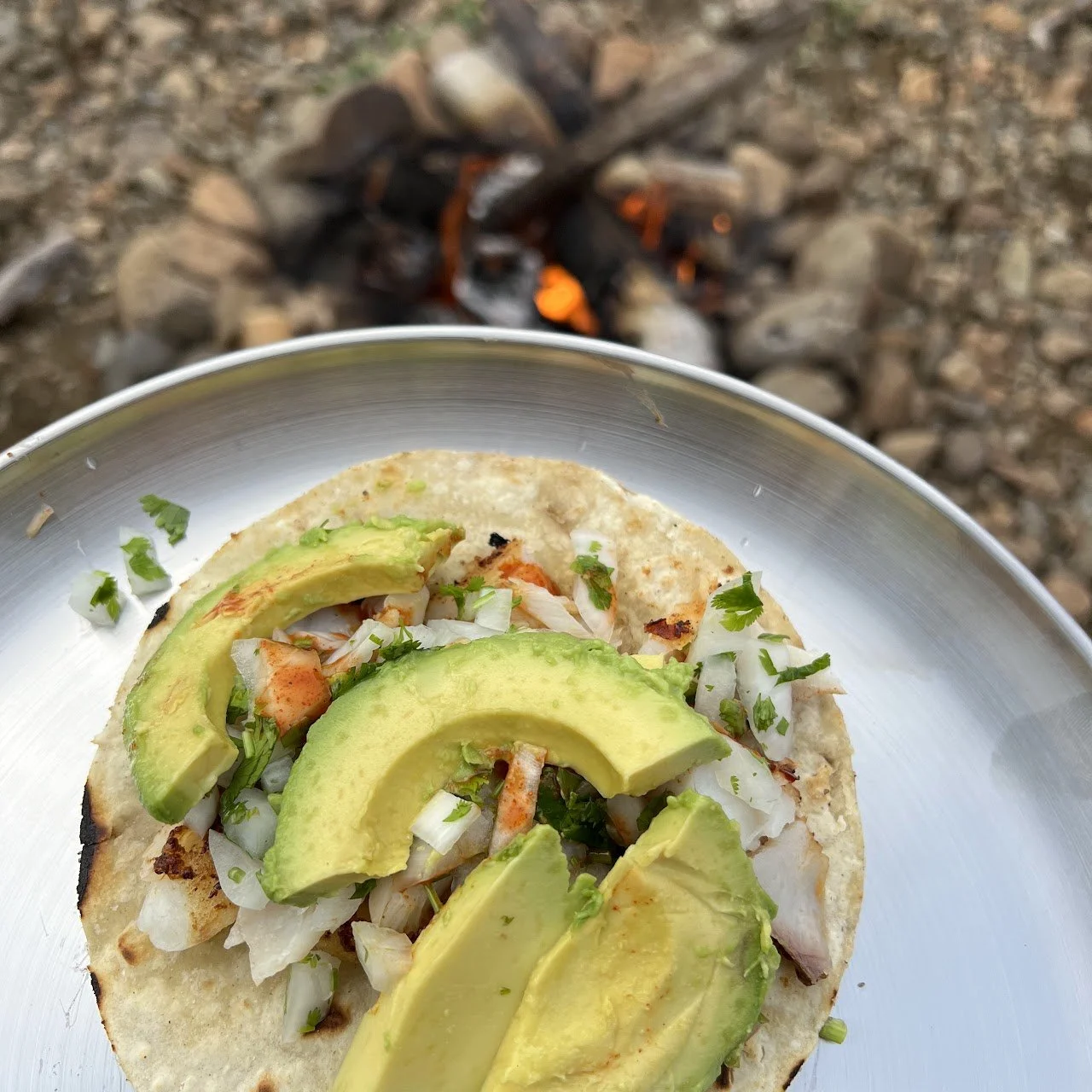 A taco with sliced avocado, chopped onions, and cilantro on a metal plate, with a campfire in the background.