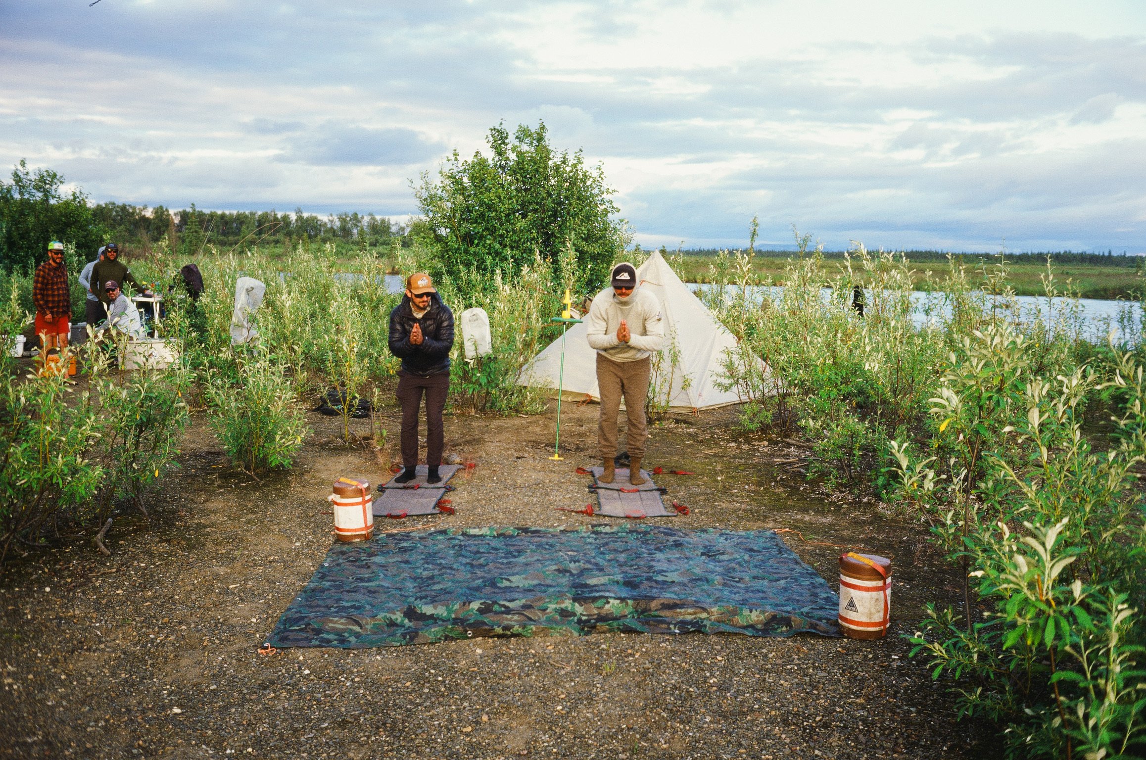 A group of people practicing yoga outdoors on a scenic riverbank, with a teepee tent and a large tree in the background, under a cloudy sky.