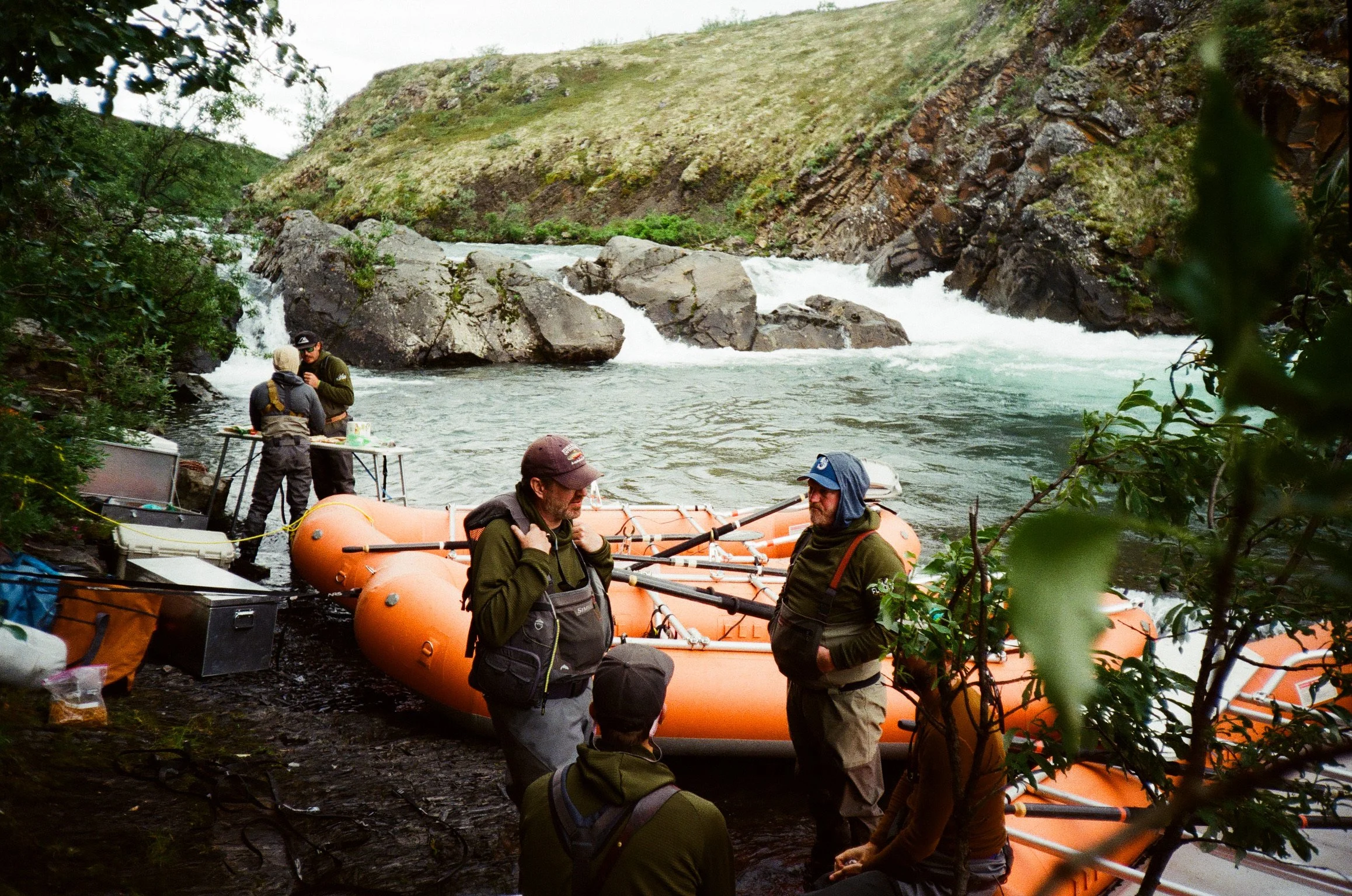 People preparing for a rafting trip on a river surrounded by green hills and rocks.