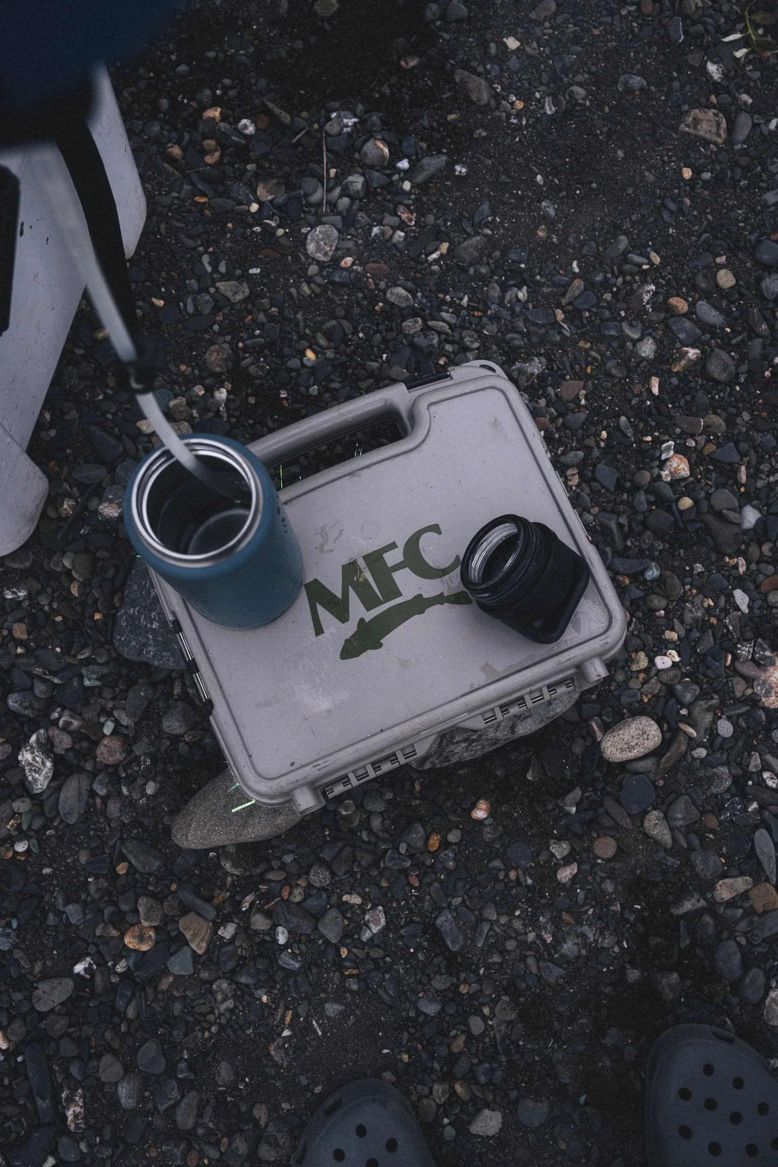 A cooler on rocky ground with an open drink container, a jar, and part of a person’s shoe visible.
