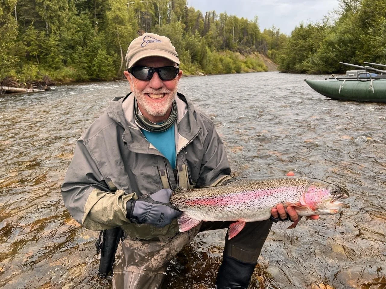 A man wearing gray fishing gear, sunglasses, and a hat, smiling while holding a large rainbow trout he caught in a river with green trees and a boat in the background.