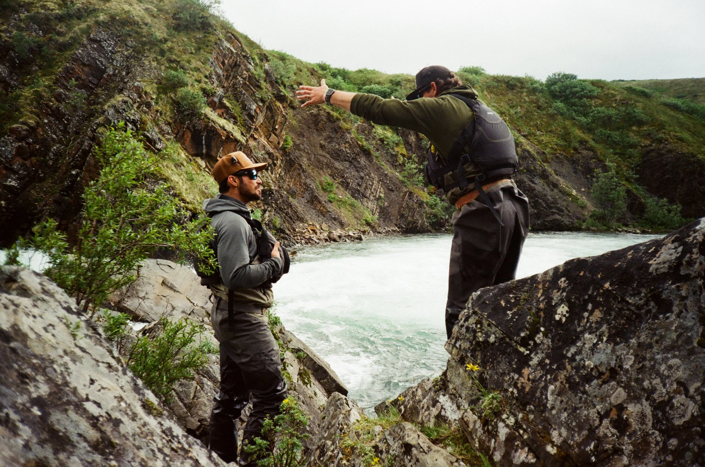 Two men outdoors by a rocky riverbank, one standing and gesturing with arms outstretched, the other sitting and listening, amidst green hills and cloudy sky.
