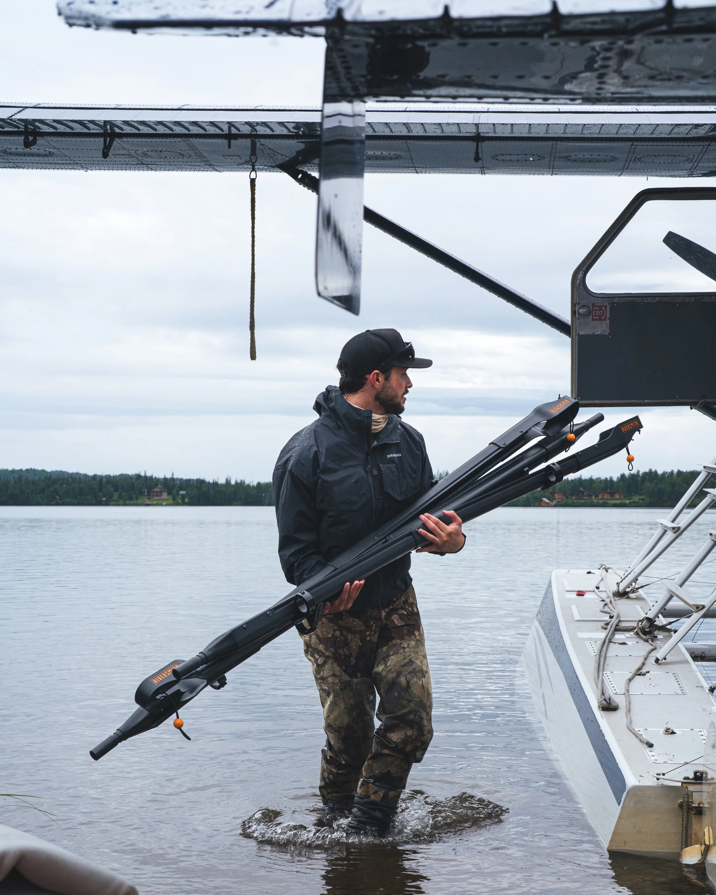 A man is in a lake or river, holding a black boat launch ramp, while standing in water up to his calves, near a dock. He is dressed in a black waterproof jacket and camouflage pants, wearing a black cap and glasses, with a cloudy sky and distant shoreline in the background.
