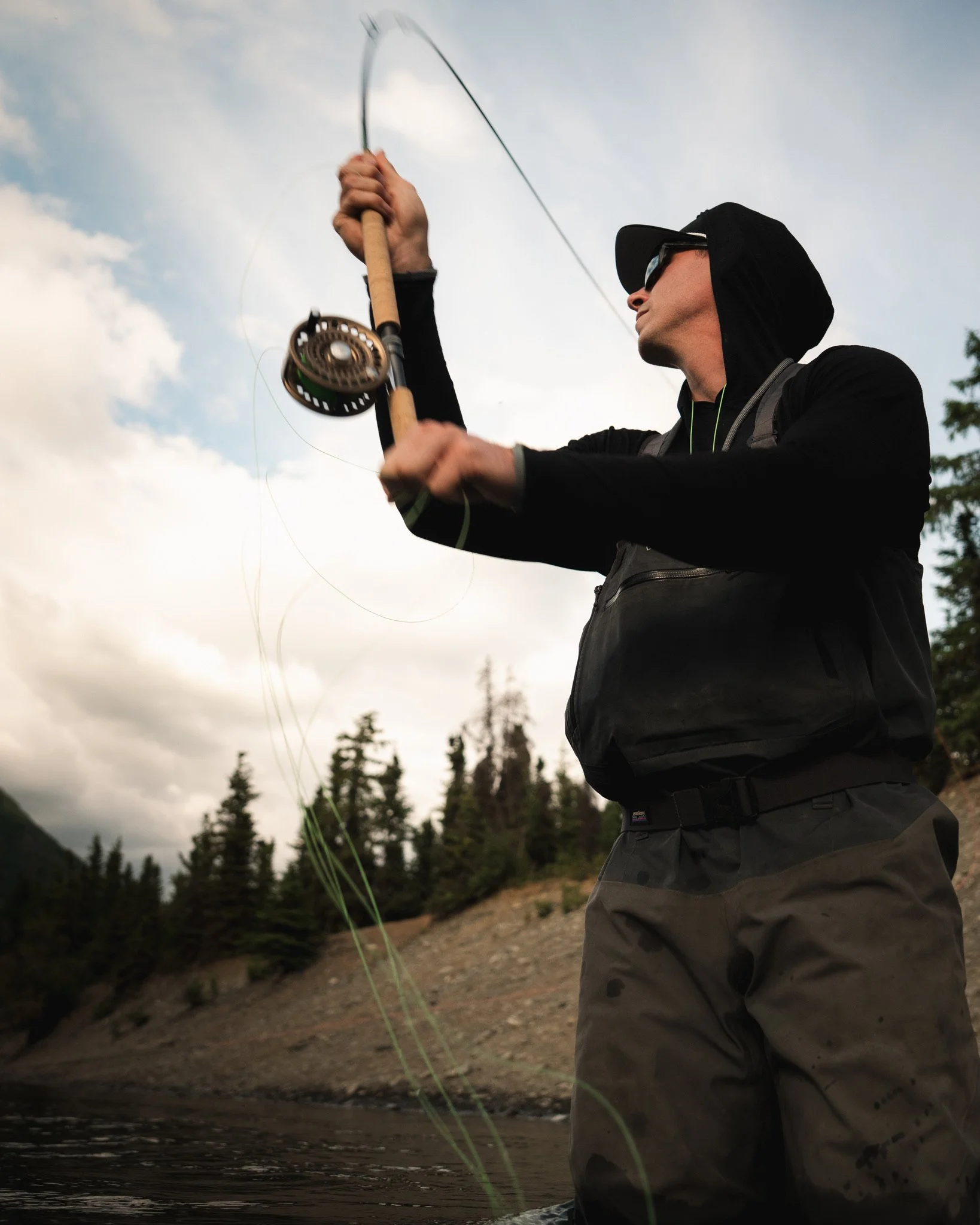 A person wearing sunglasses, a black hoodie, and gray pants is fly fishing in a river with a scenic backdrop of trees and mountains under a cloudy sky.