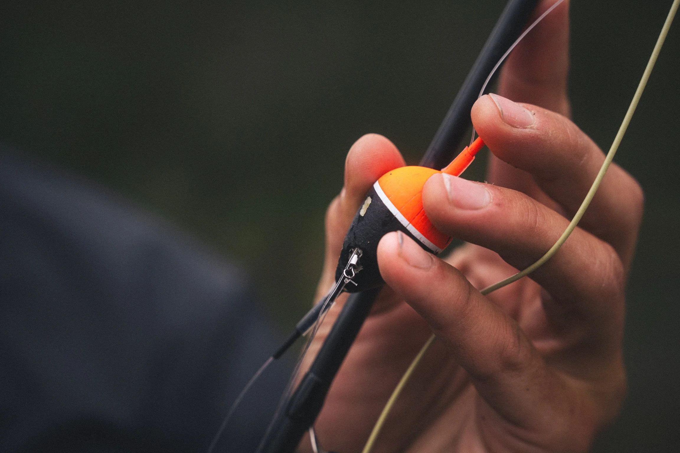 Close-up of a person's hand holding a fishing rod with a bright orange float, over a dark background.