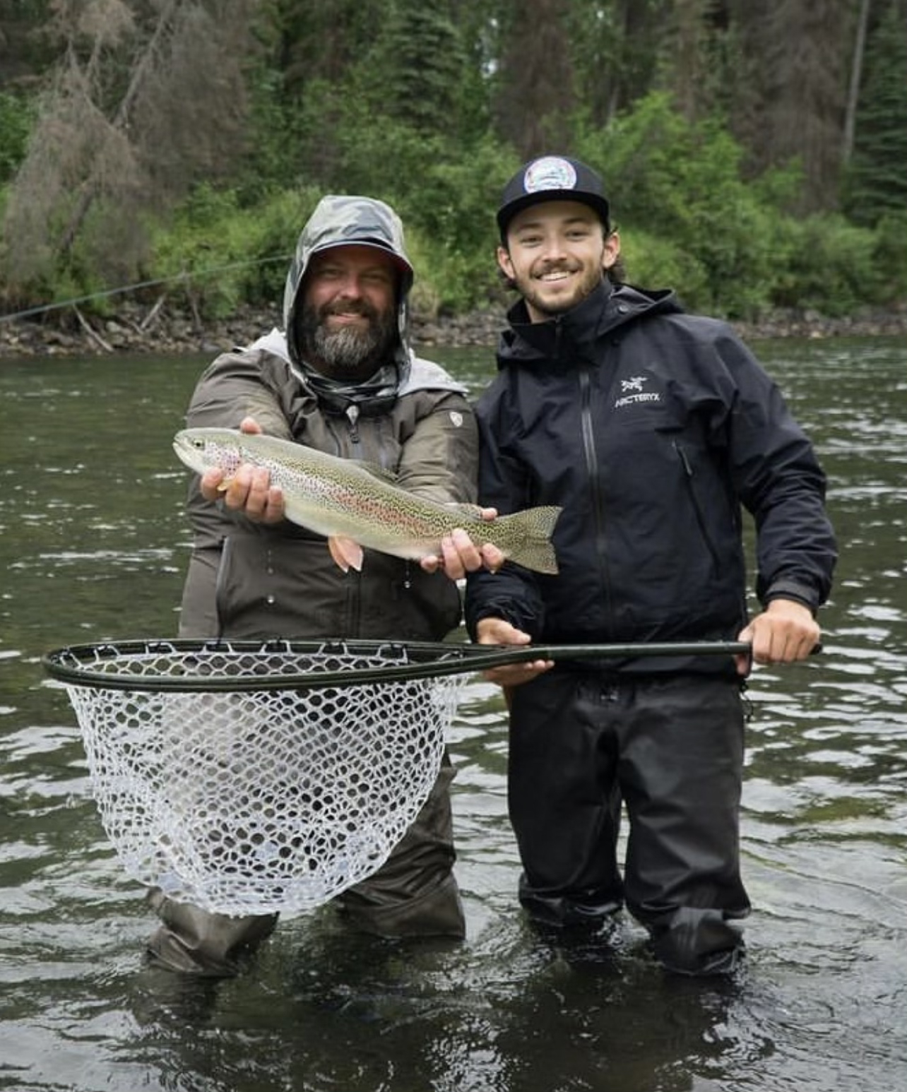 Two men stand in a river holding a large rainbow trout, with one man holding a fishing net, surrounded by trees.