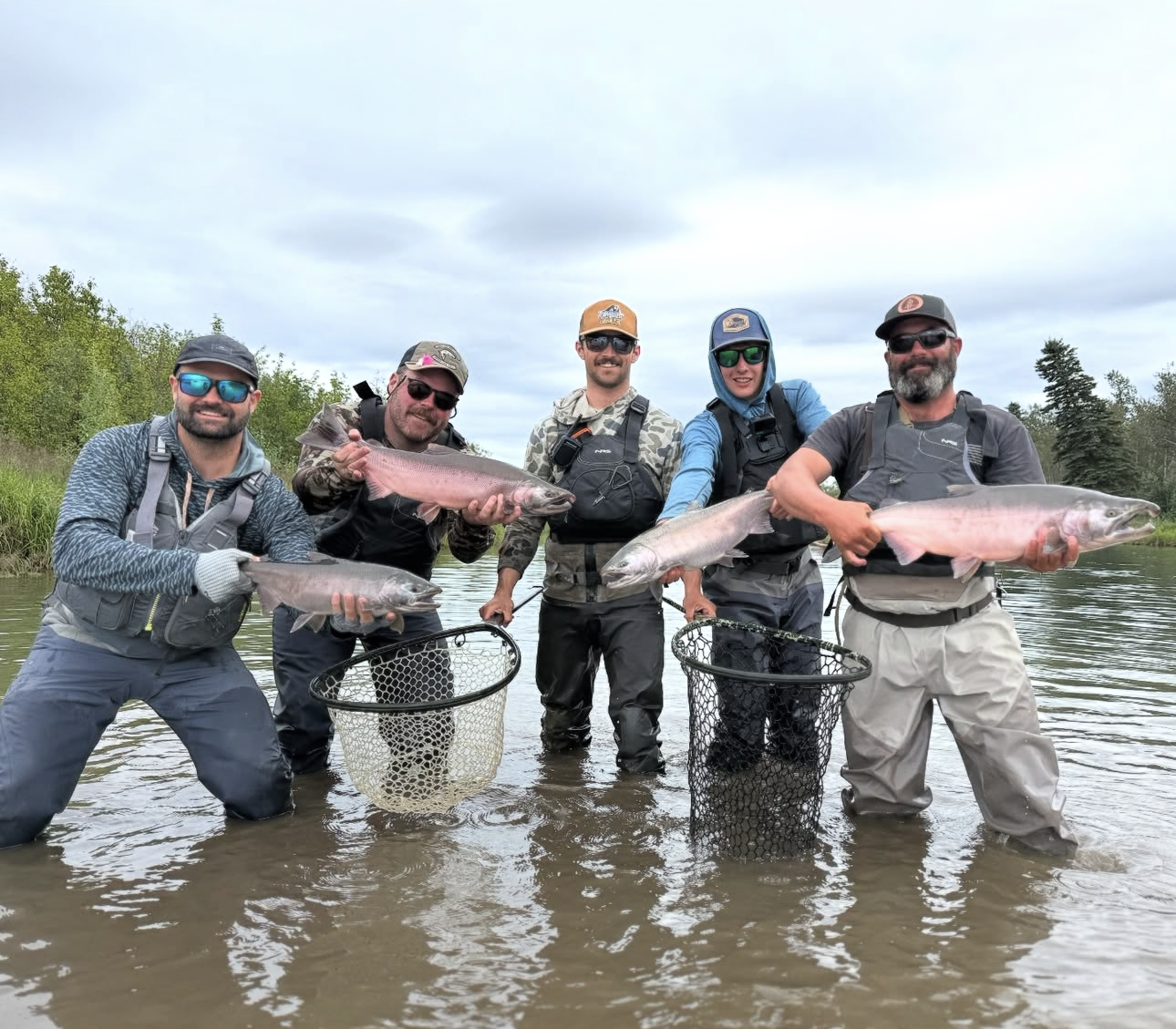 Five men fishing and holding large fish in a river with green trees in the background.