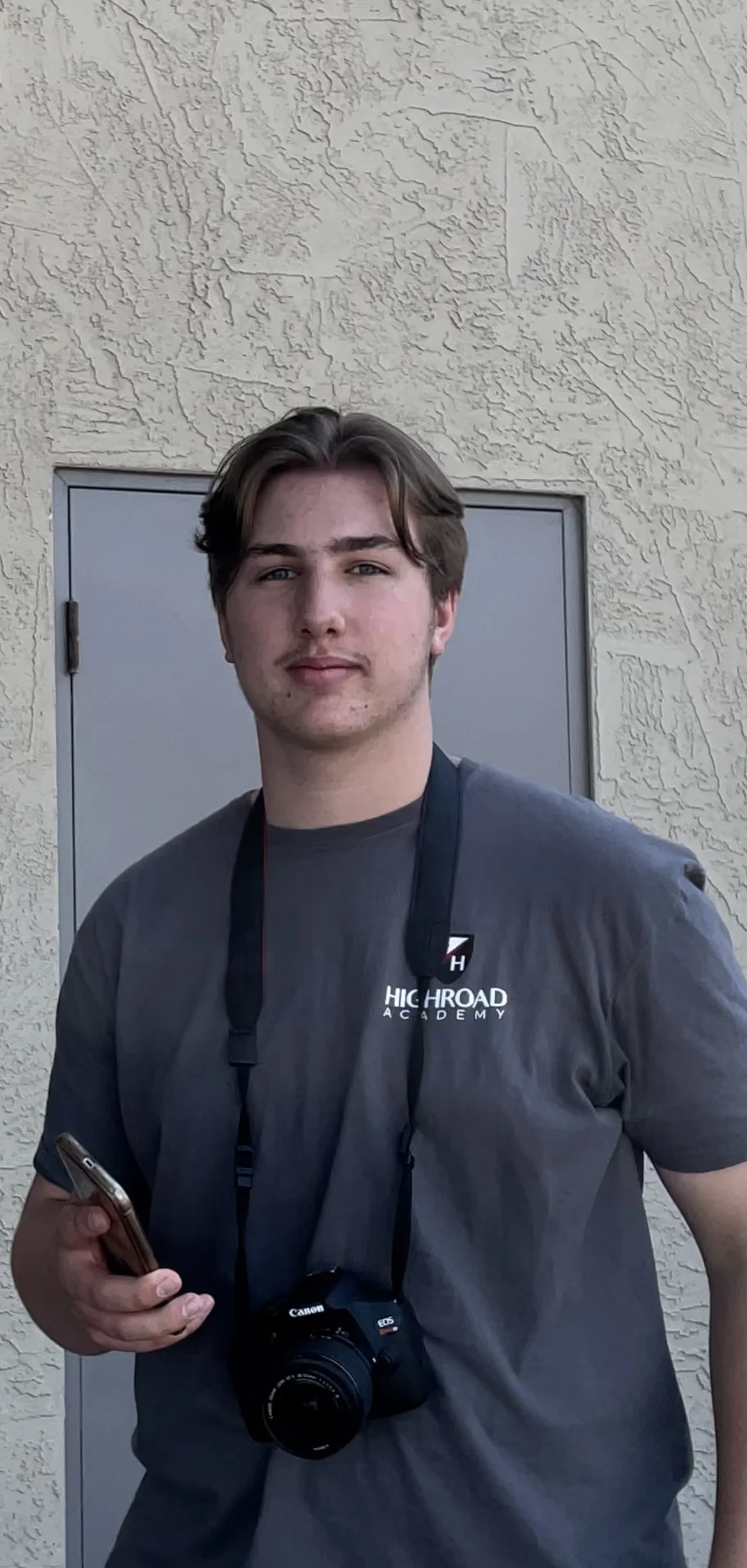 Young man with brown hair wearing a gray t-shirt with 'High Road Academy' logo, holding a smartphone and a Canon camera around his neck, standing against a textured beige wall and gray door.