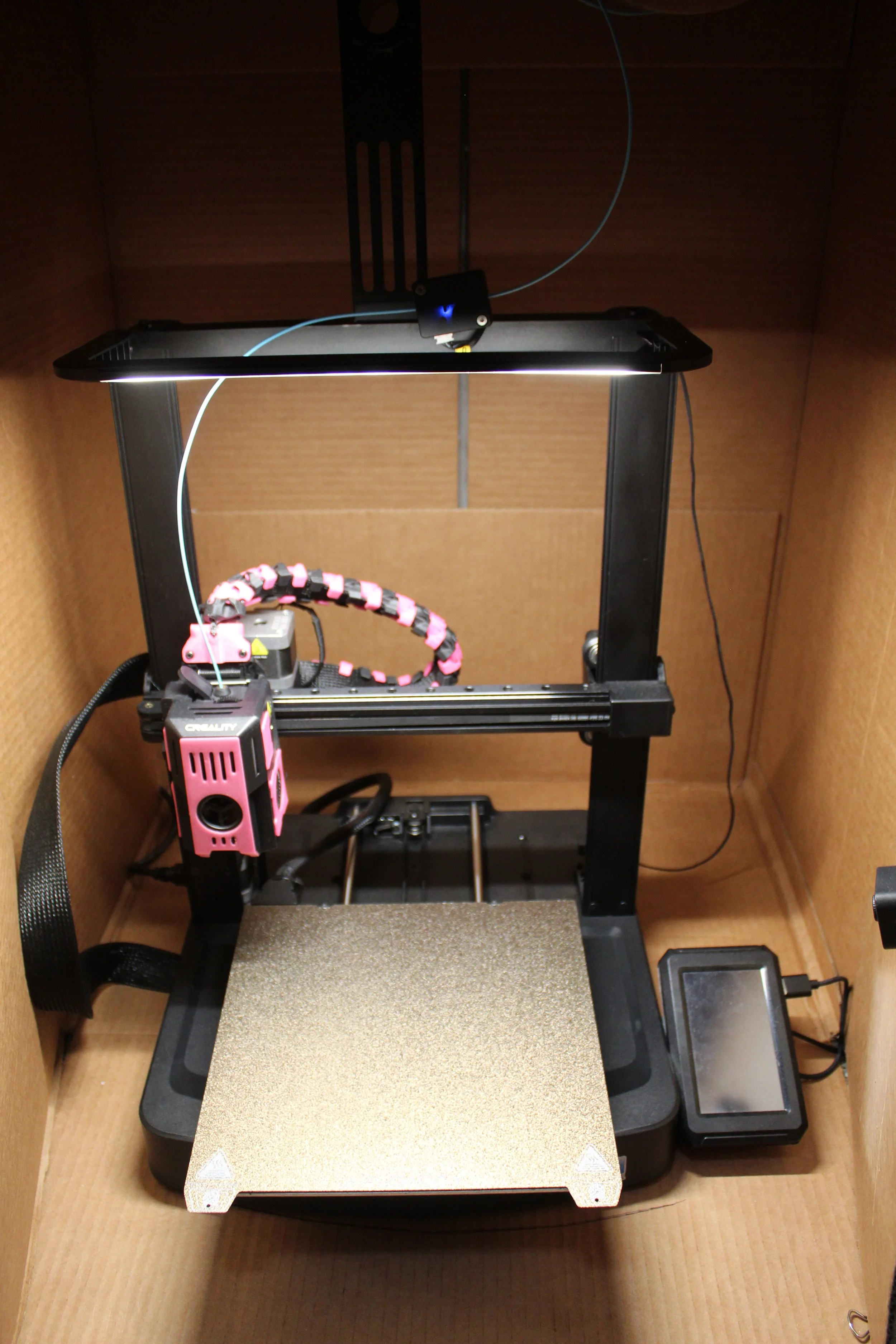 A 3D printer inside a cardboard enclosure with a heated bed, filament, and lighting.