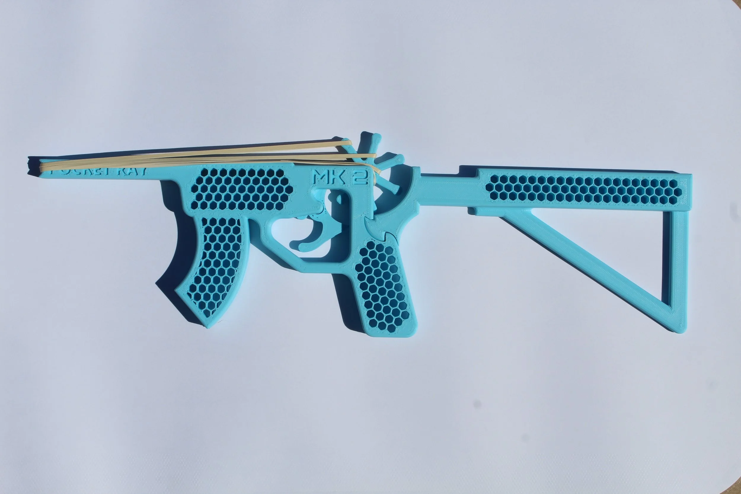 Light blue 3D-printed toy gun with a honeycomb pattern, labeled 'MK 2', and rubber bands attached on top, set against a white background.