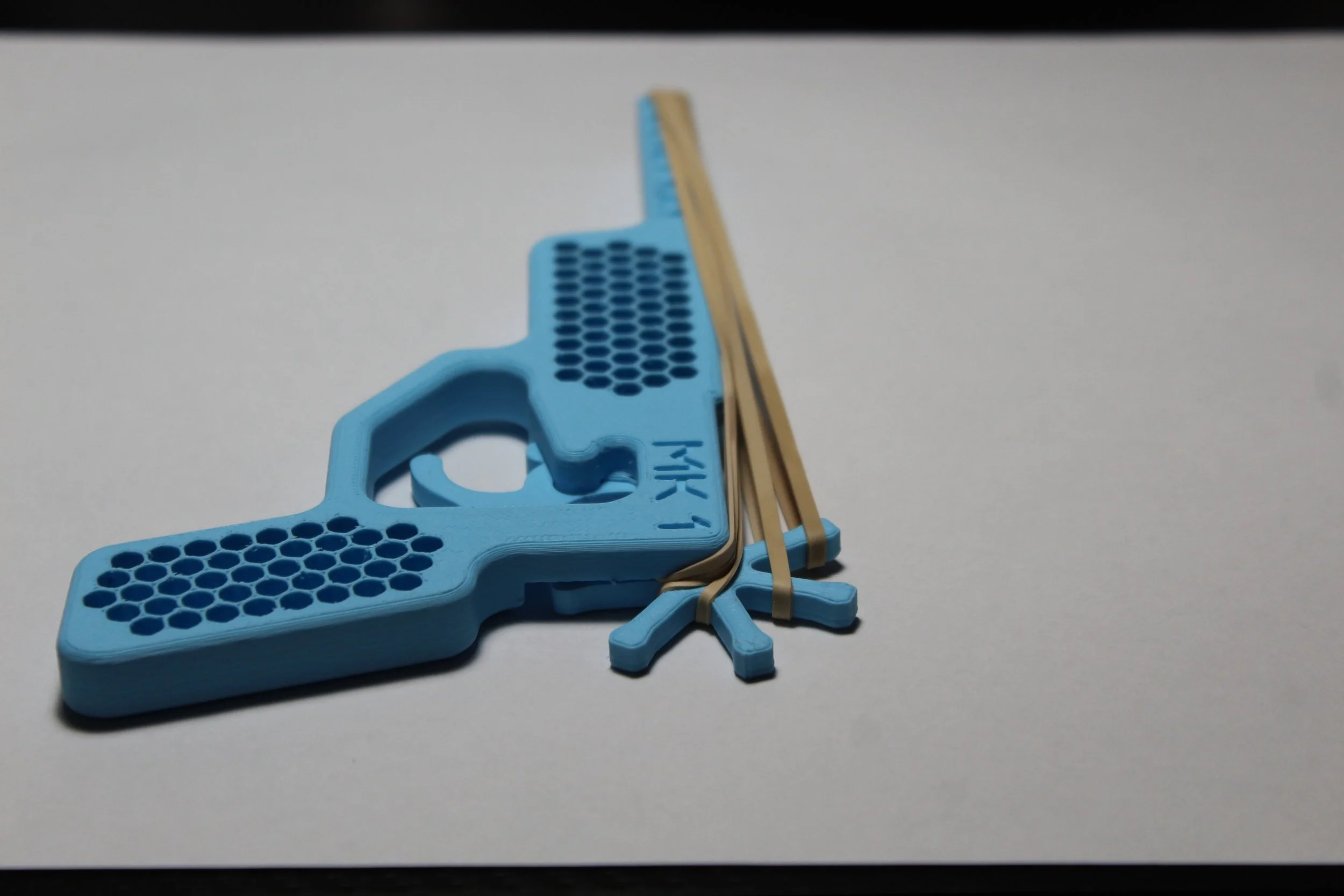 3D-printed blue gun-shaped object with rubber bands wrapped around it, placed on a white surface.