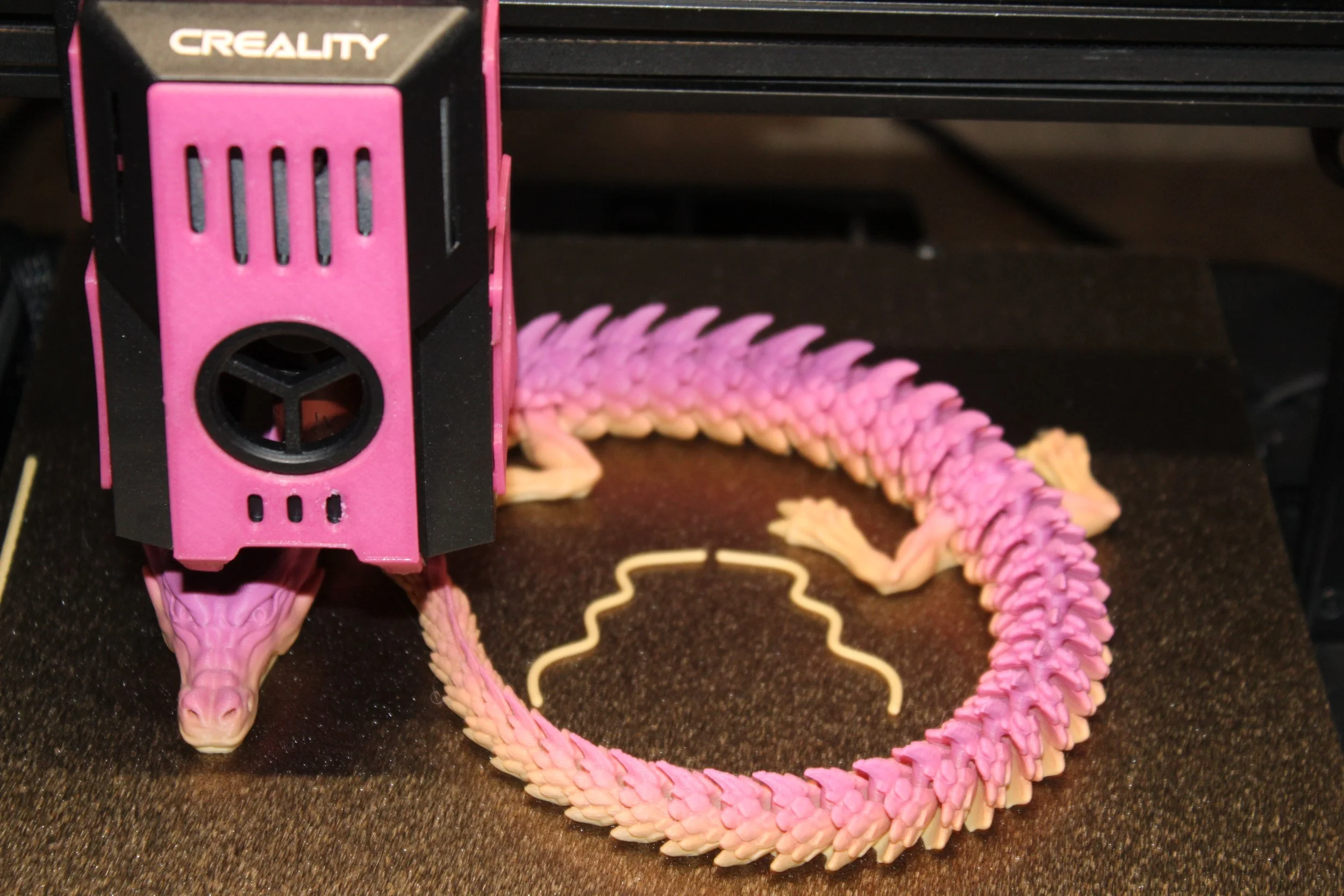 Pink and black 3D printer printing a pink and peach and yellow flexible dragon