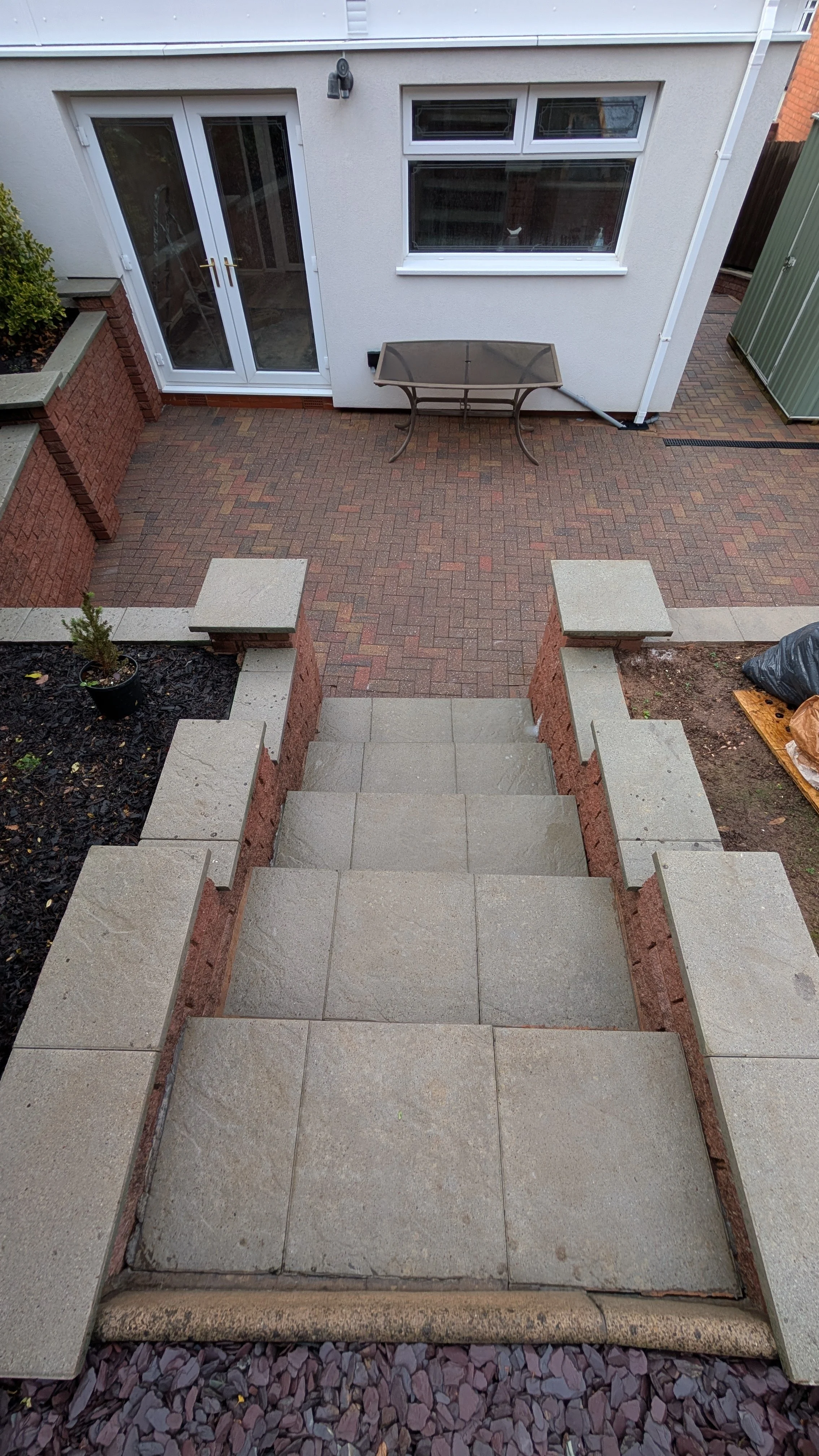 Patio cleaning in Marlow. View from the top of a short outdoor staircase leading to a backyard patio area with a brick and stone paved surface, a glass patio table, and a white house with double glass doors and windows.